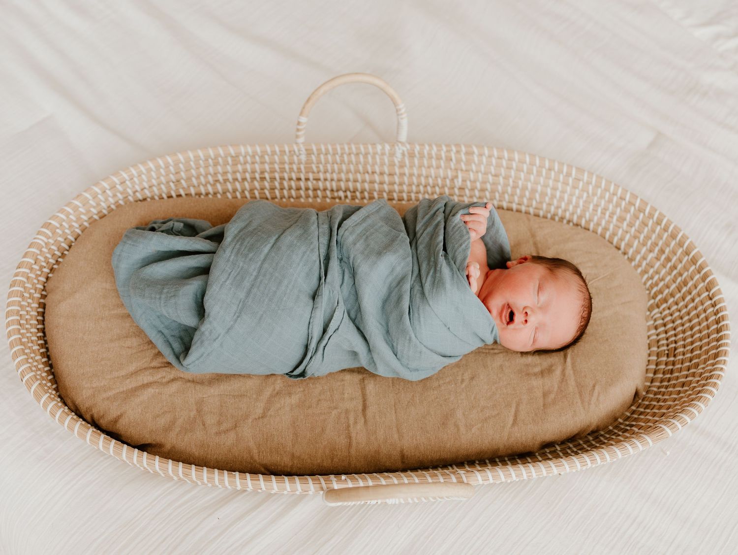 A sleeping infant wearing teal clothing lies peacefully in a round woven bassinet.