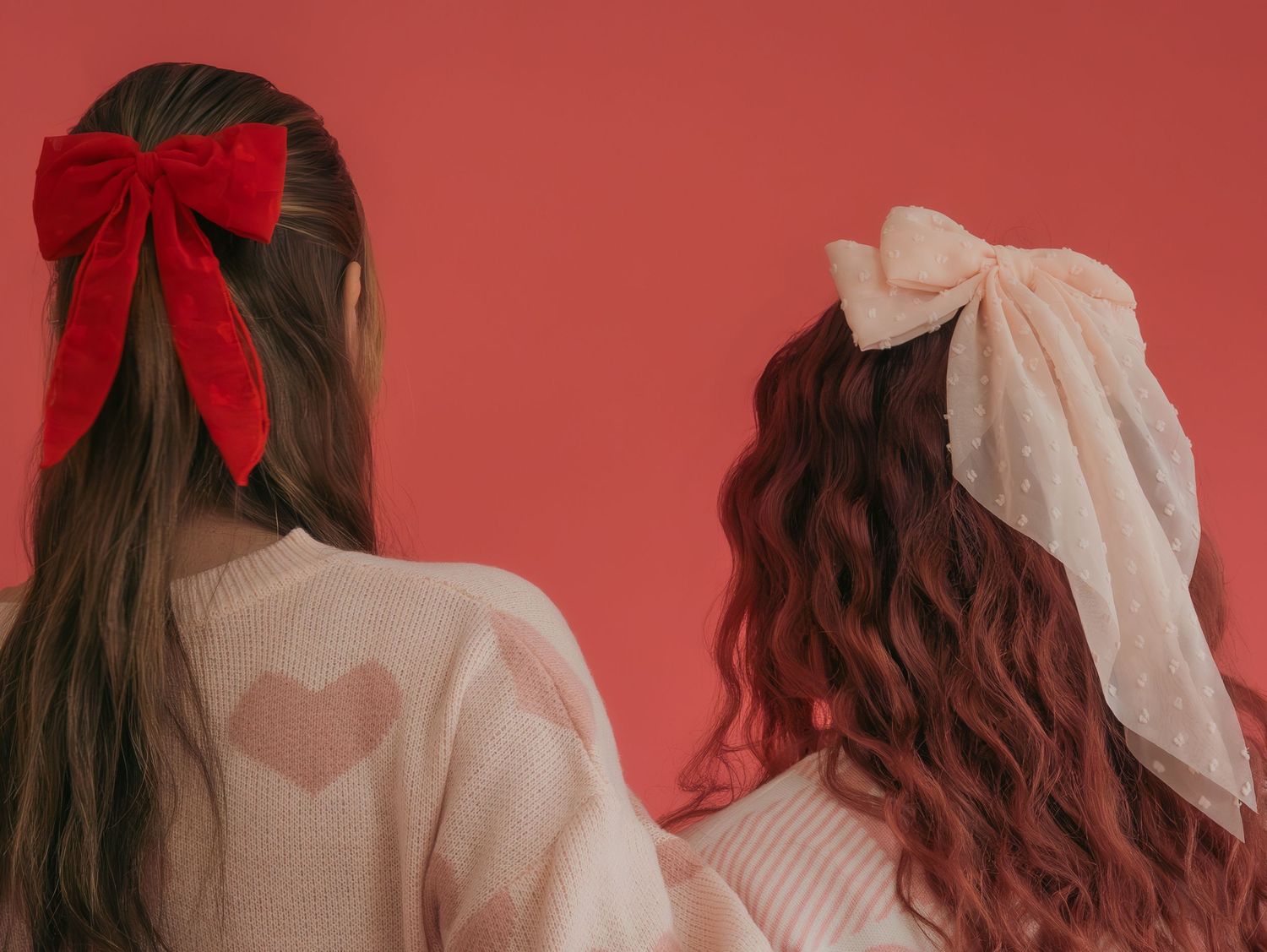 Back view of two people wearing pink sweaters with hair bows against coral background.