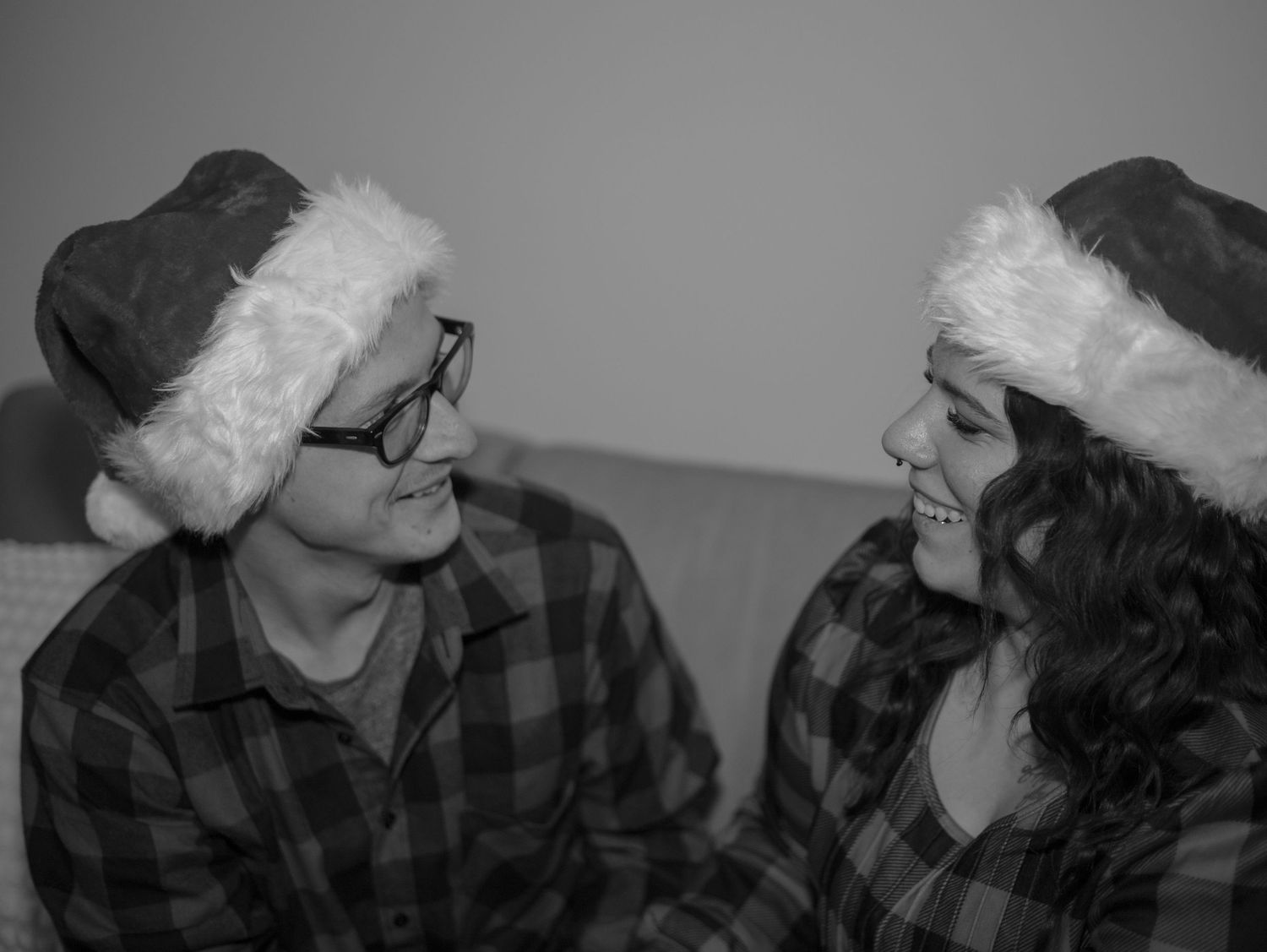 A black and white portrait of two people in plaid shirts and Santa hats sharing a moment.