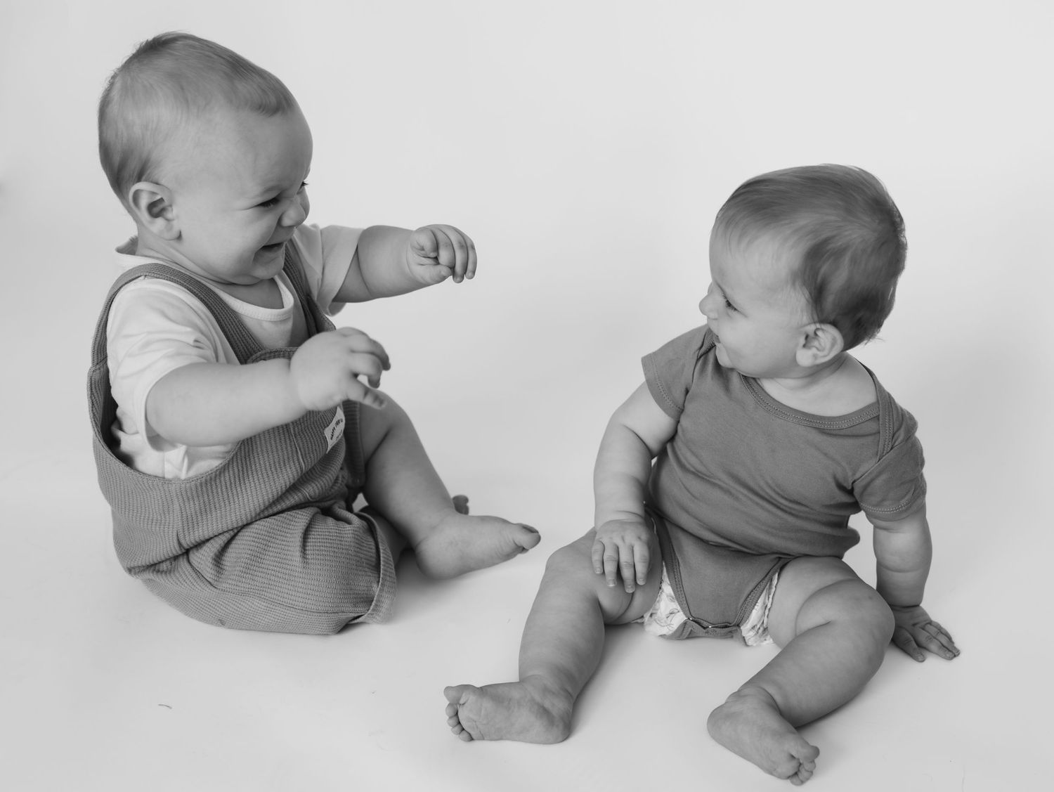 Black and white vintage photo shows two seated babies in onesies looking at each other while exploring.