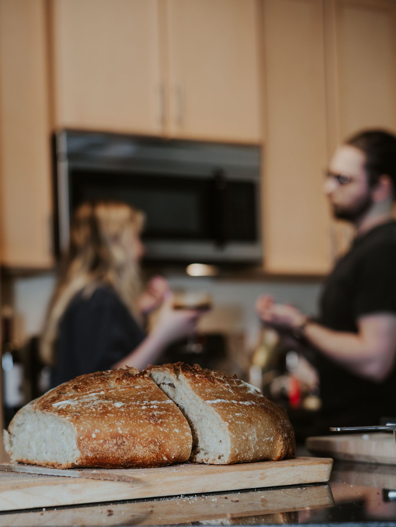 A freshly baked rustic sourdough bread loaf being sliced on a wooden cutting board in a kitchen setting.