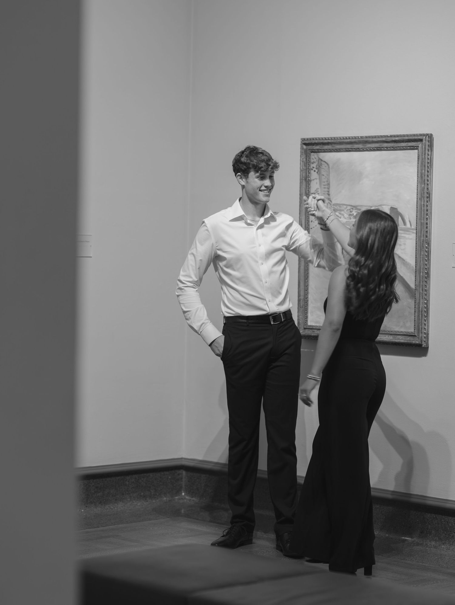 A couple in formal attire stands in an art gallery, with the woman gesturing to a framed artwork on the wall.