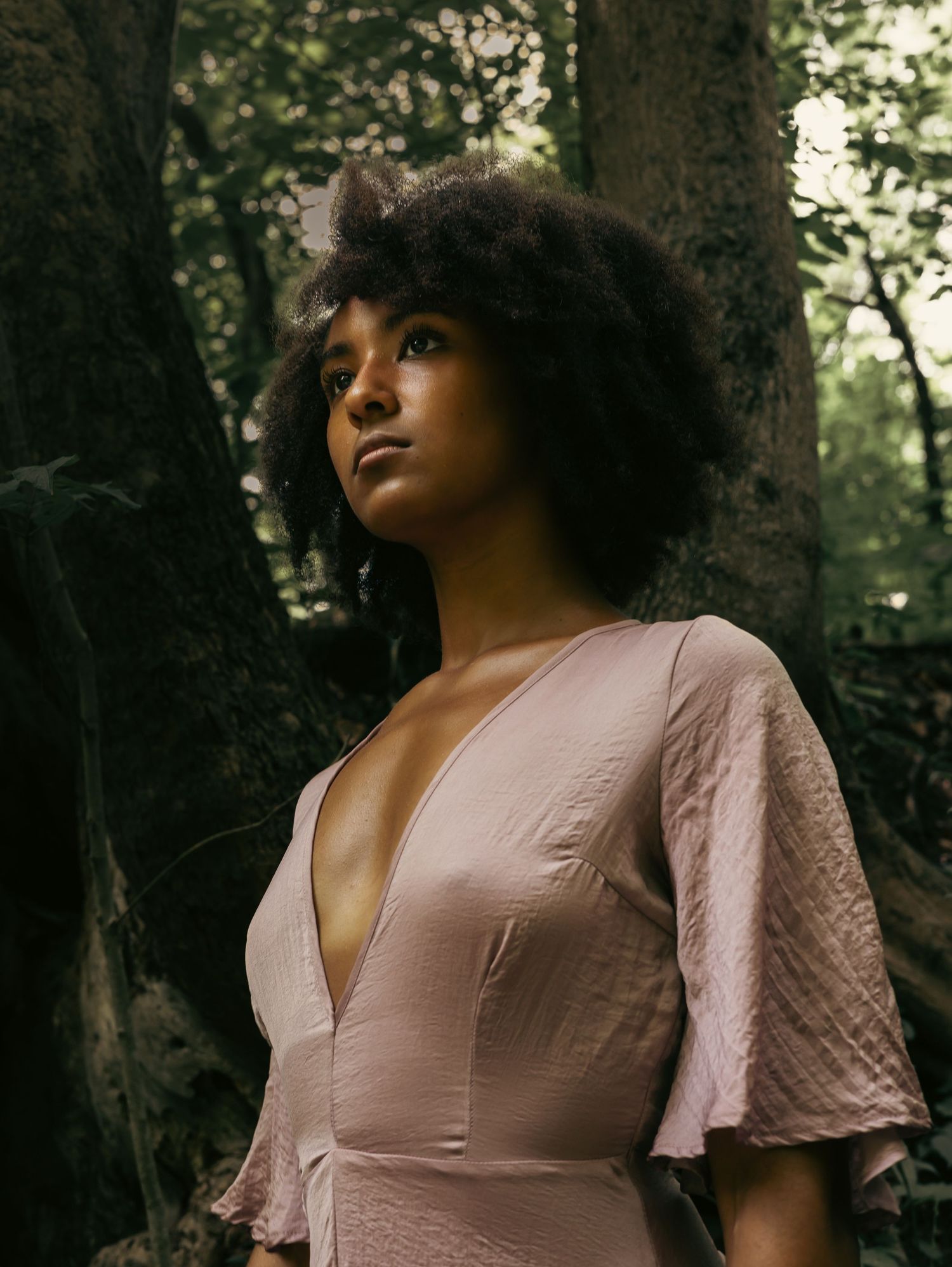 A person in a pink dress poses thoughtfully against a dark wooded forest background in natural lighting.