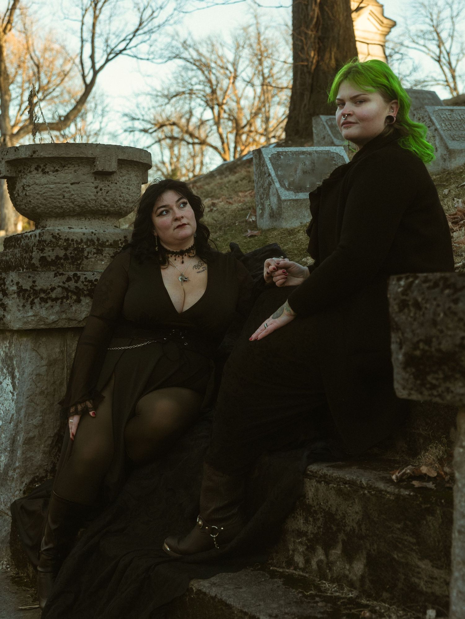 Two people in dark gothic attire relax on stone steps in a cemetery during autumn.
