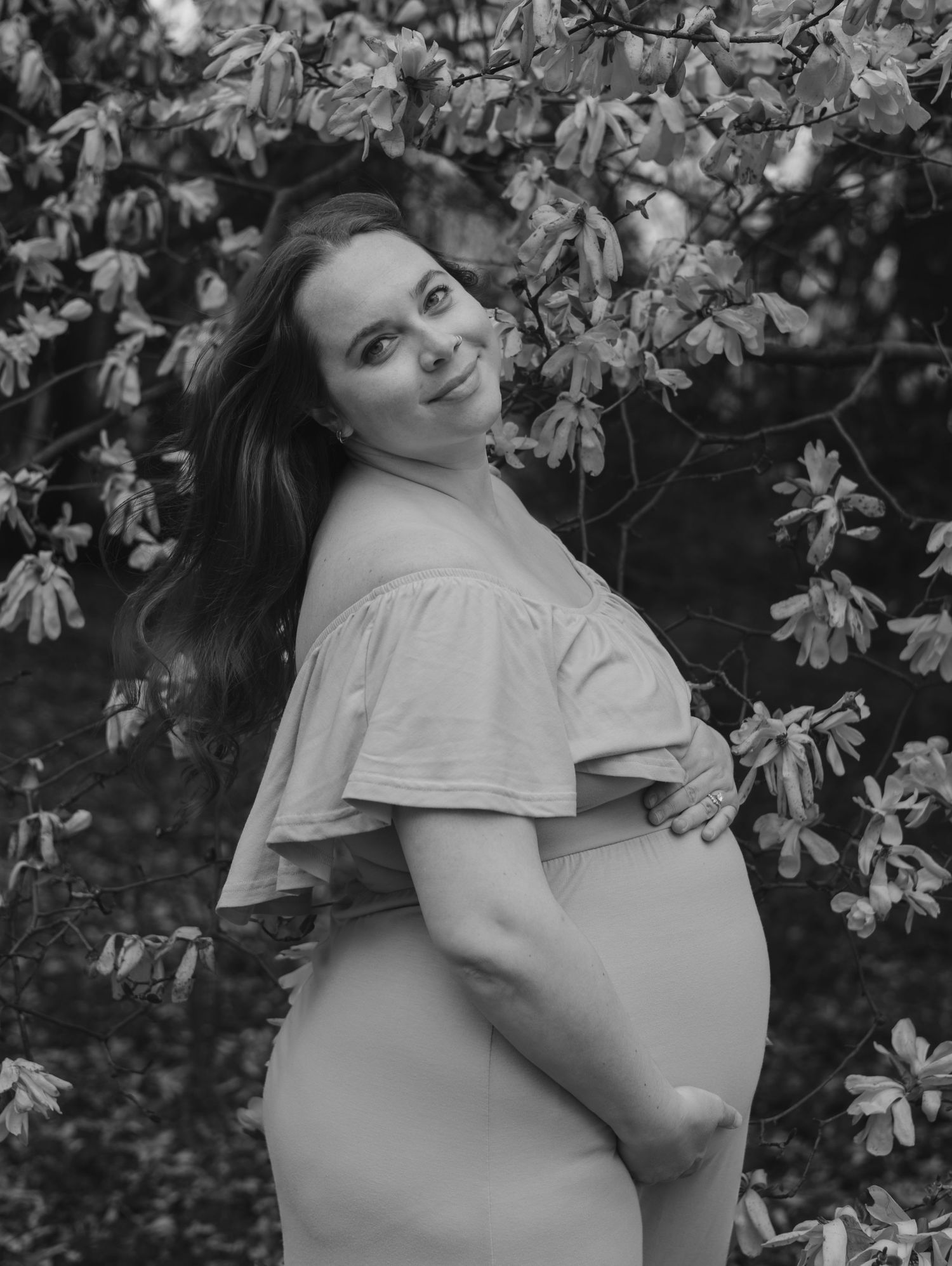 A black and white maternity photoshoot showcases an expectant mother posing gracefully among flowering tree branches.