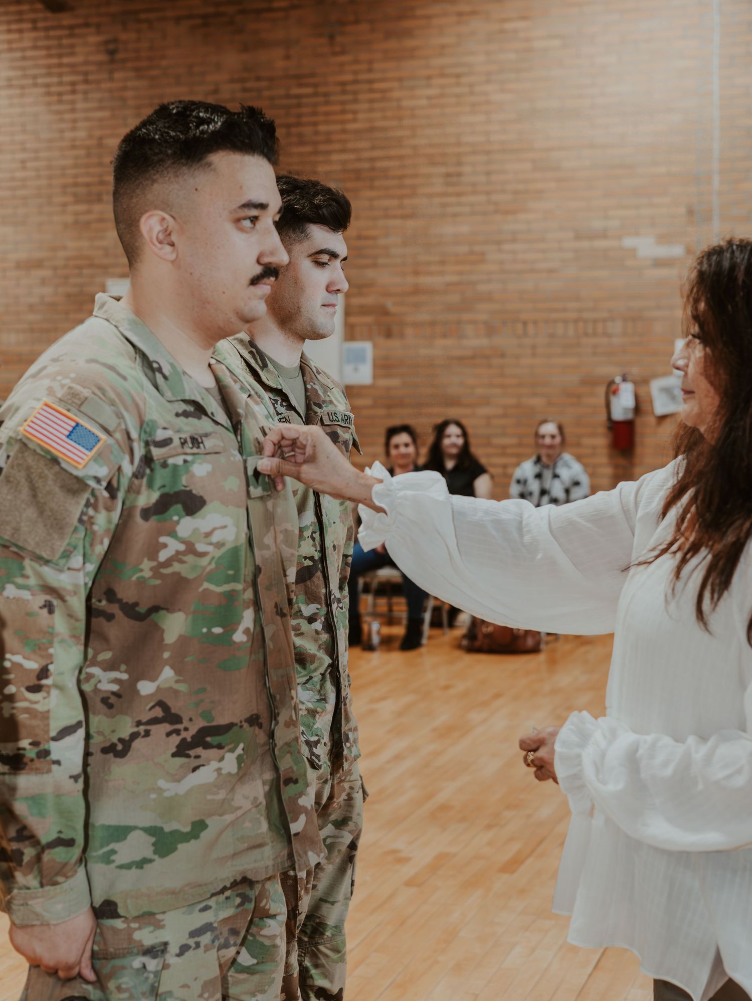 Two U.S. military service members in camouflage uniforms stand at attention while being blessed during a ceremony.