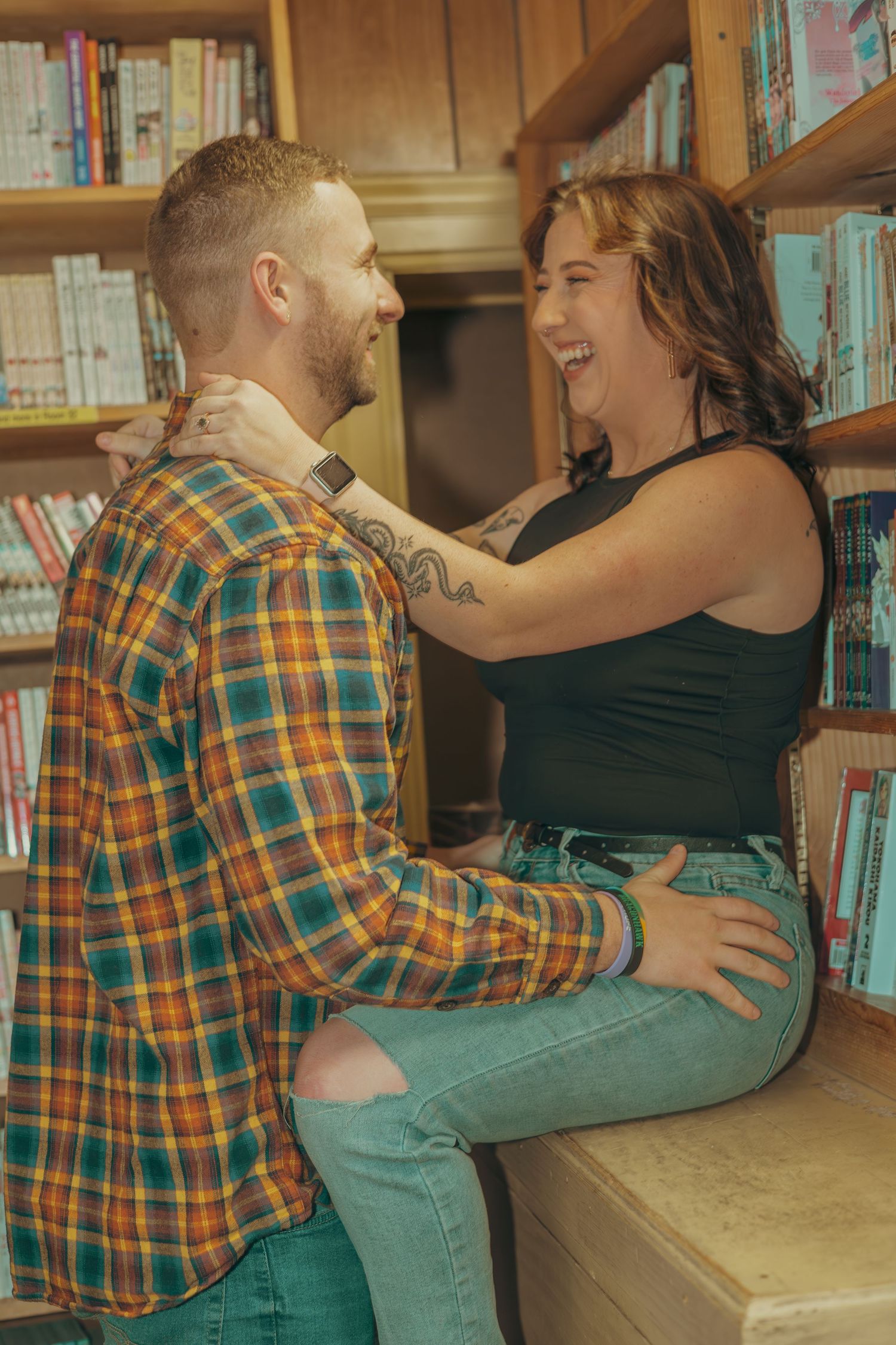 A romantic couple embraces and laughs together in a library setting with bookshelves in the background.