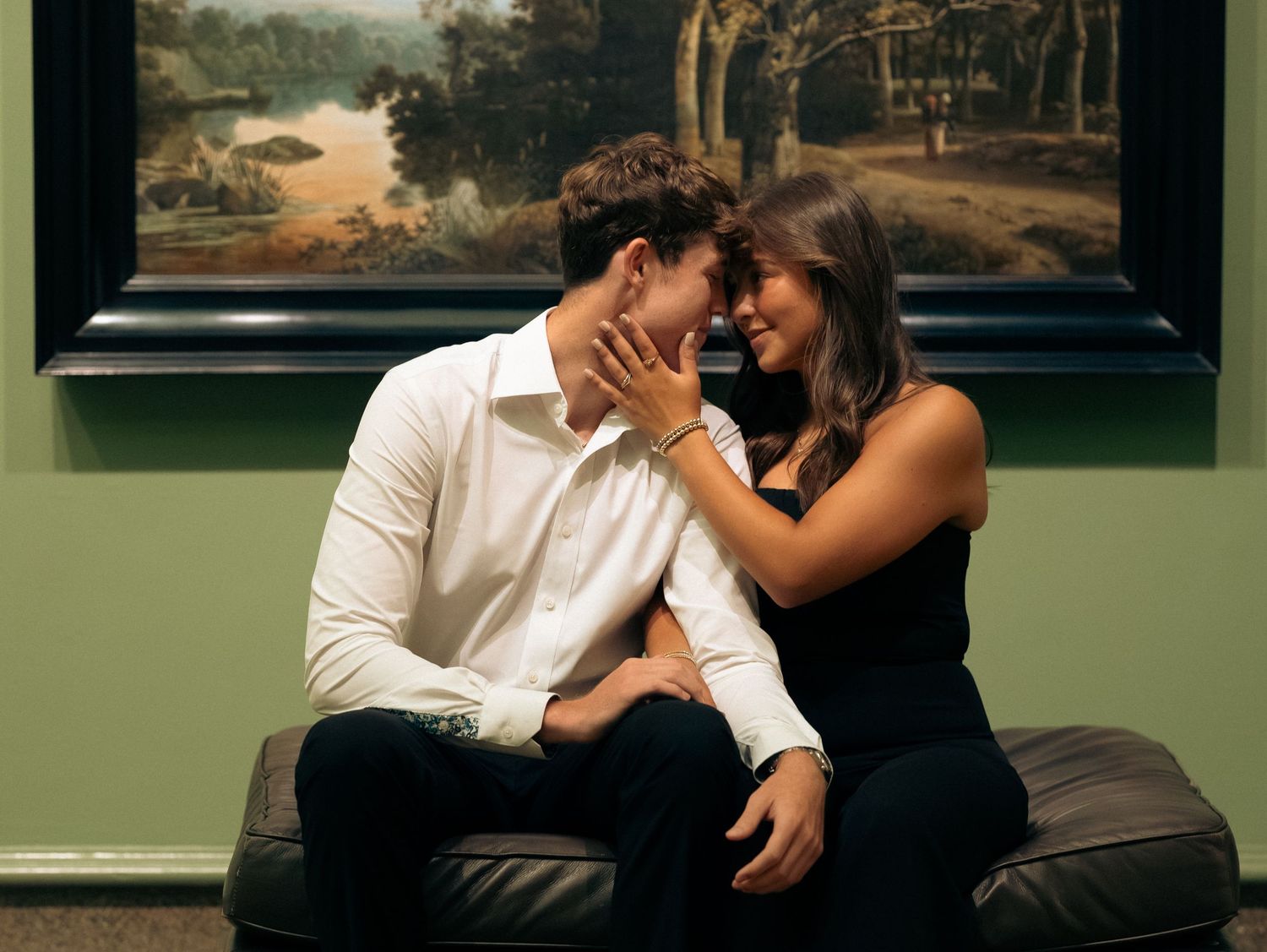 A romantic couple shares intimate moments on a brown leather couch in an art gallery or museum setting.