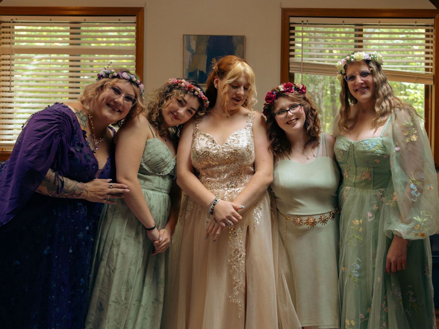 A group of five people in elegant formal dresses with floral crowns pose together at a special event.