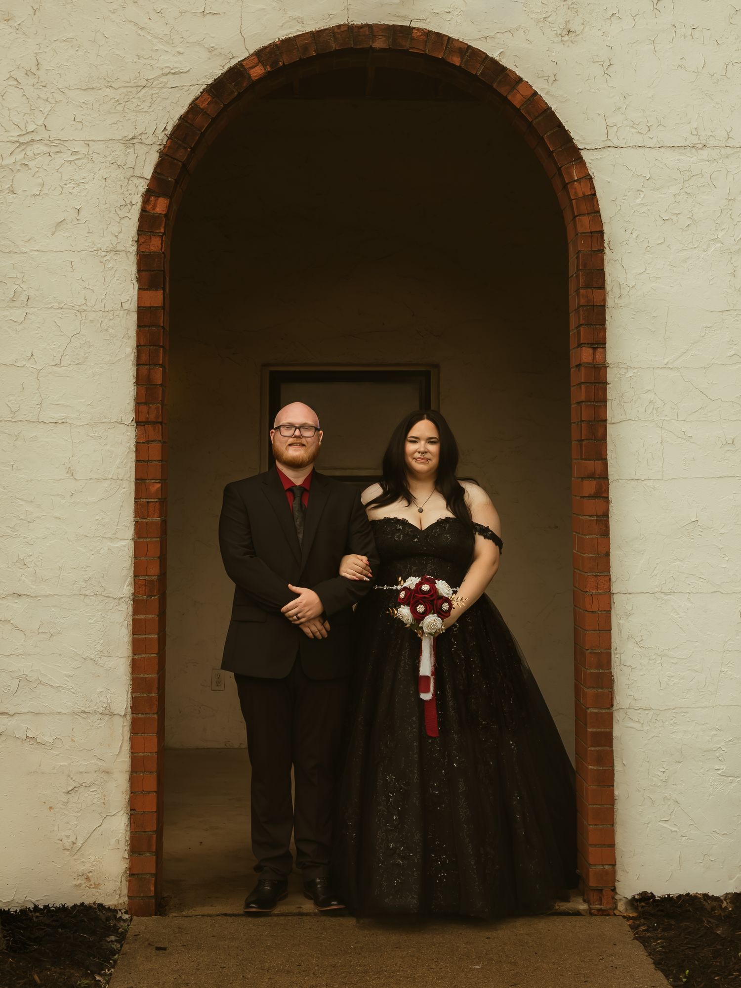 Wedding couple poses under brick archway, bride in black gown with bouquet, groom in black suit red tie.