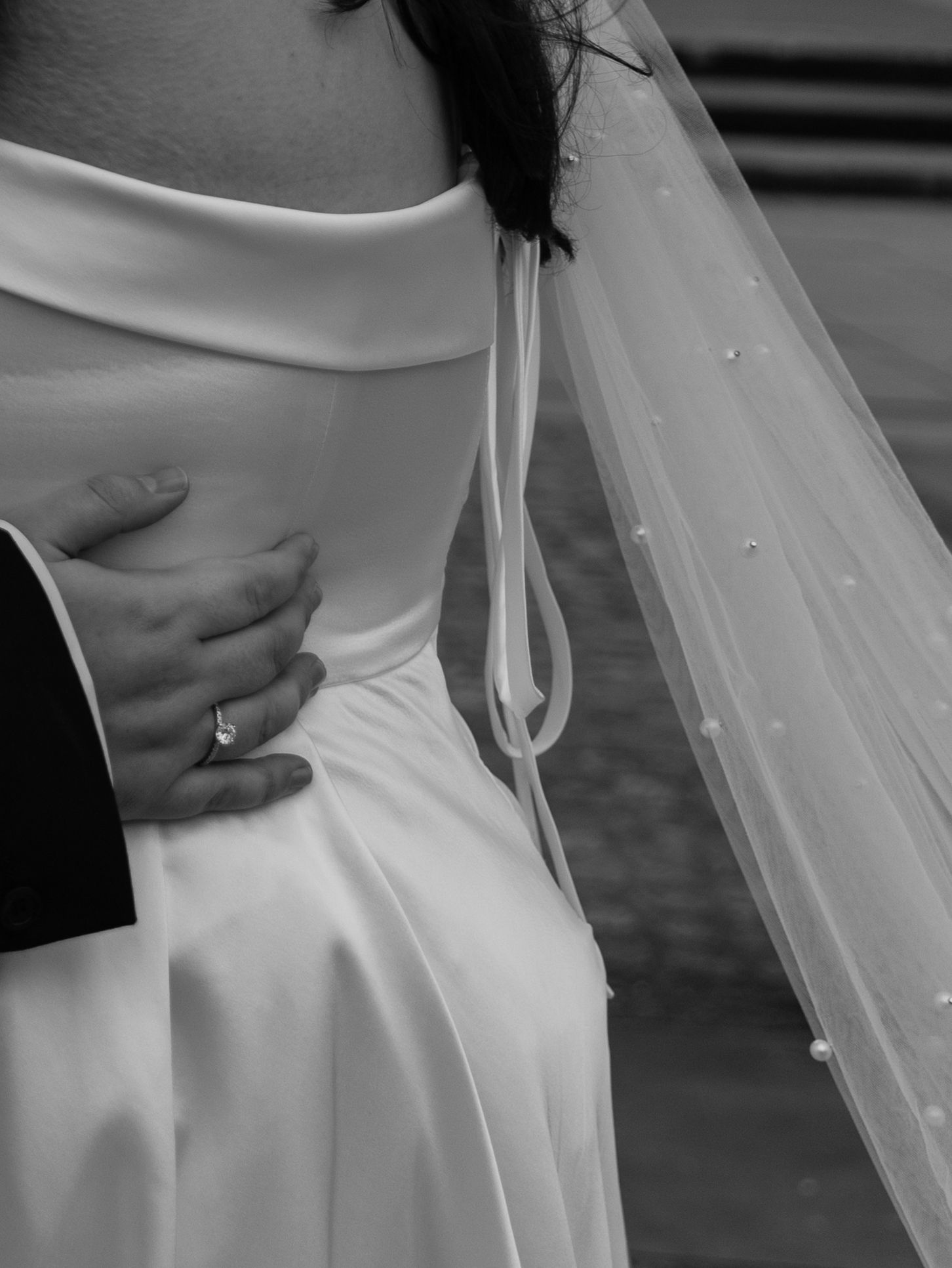 Close-up black and white photo of a wedding dress detail showing delicate veil and hand with engagement ring.
