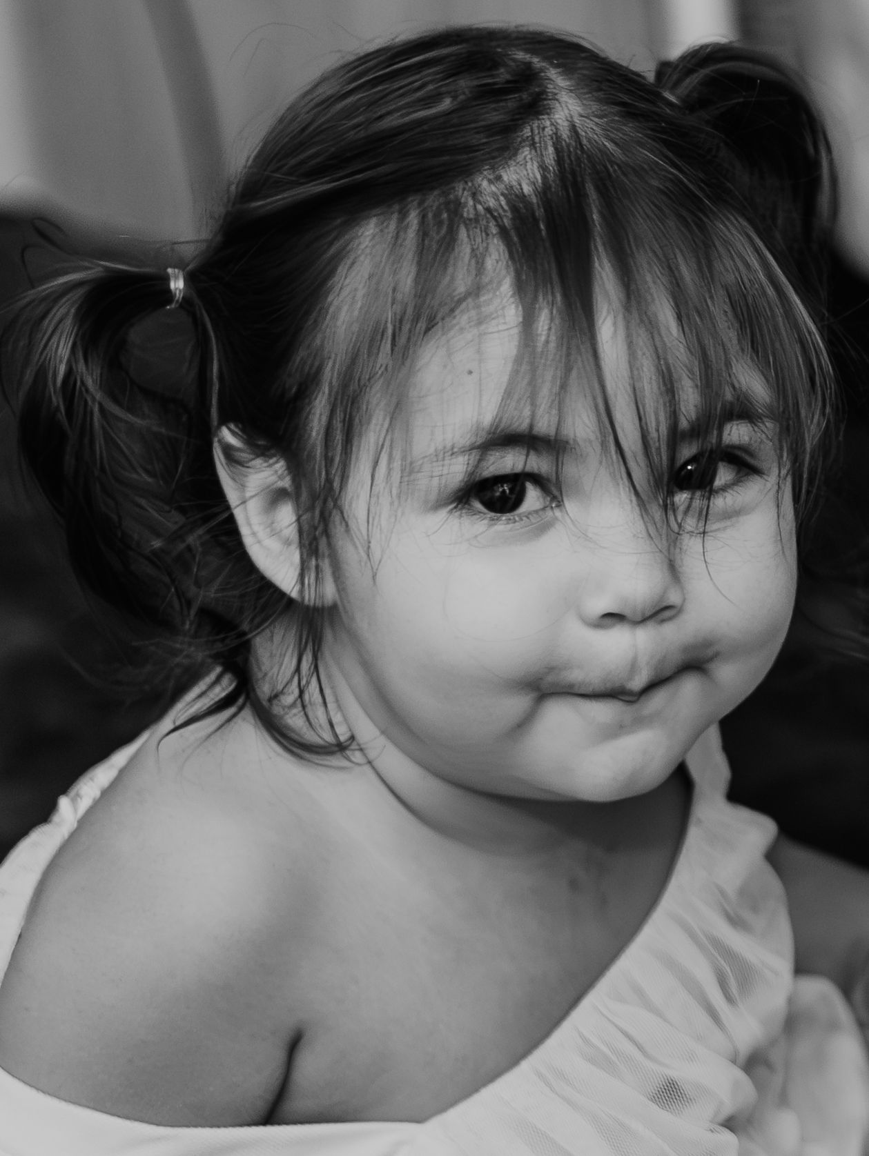 A black and white portrait of a young child with pigtails and bangs making a sweet expression in casual dress.