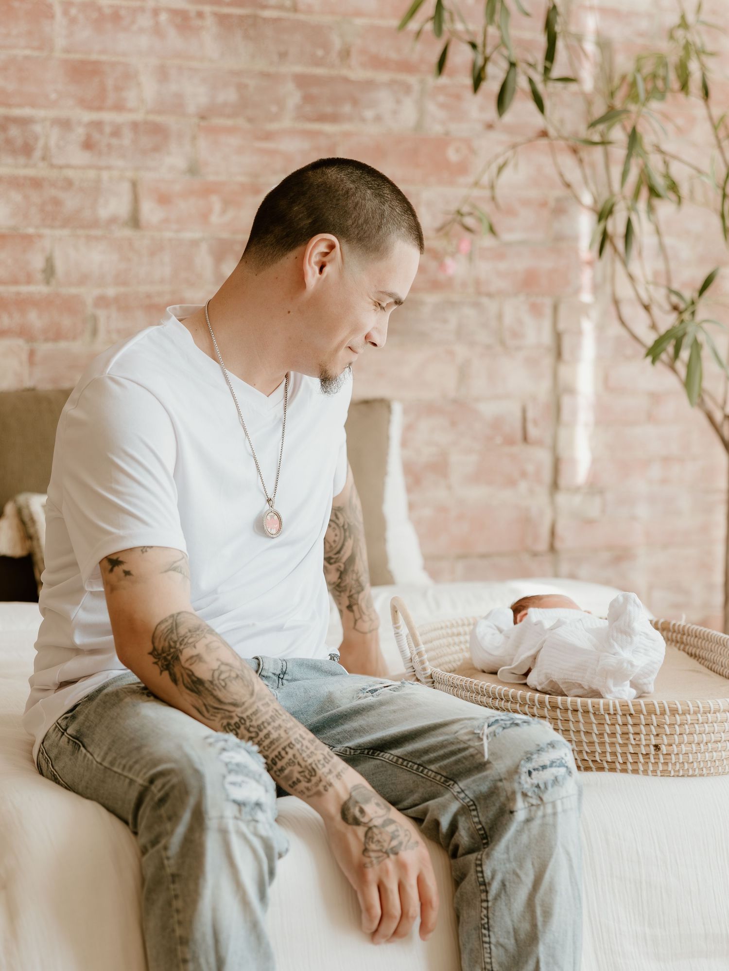 Person in white t-shirt with arm tattoos sits thoughtfully on woven furniture against brick wall.