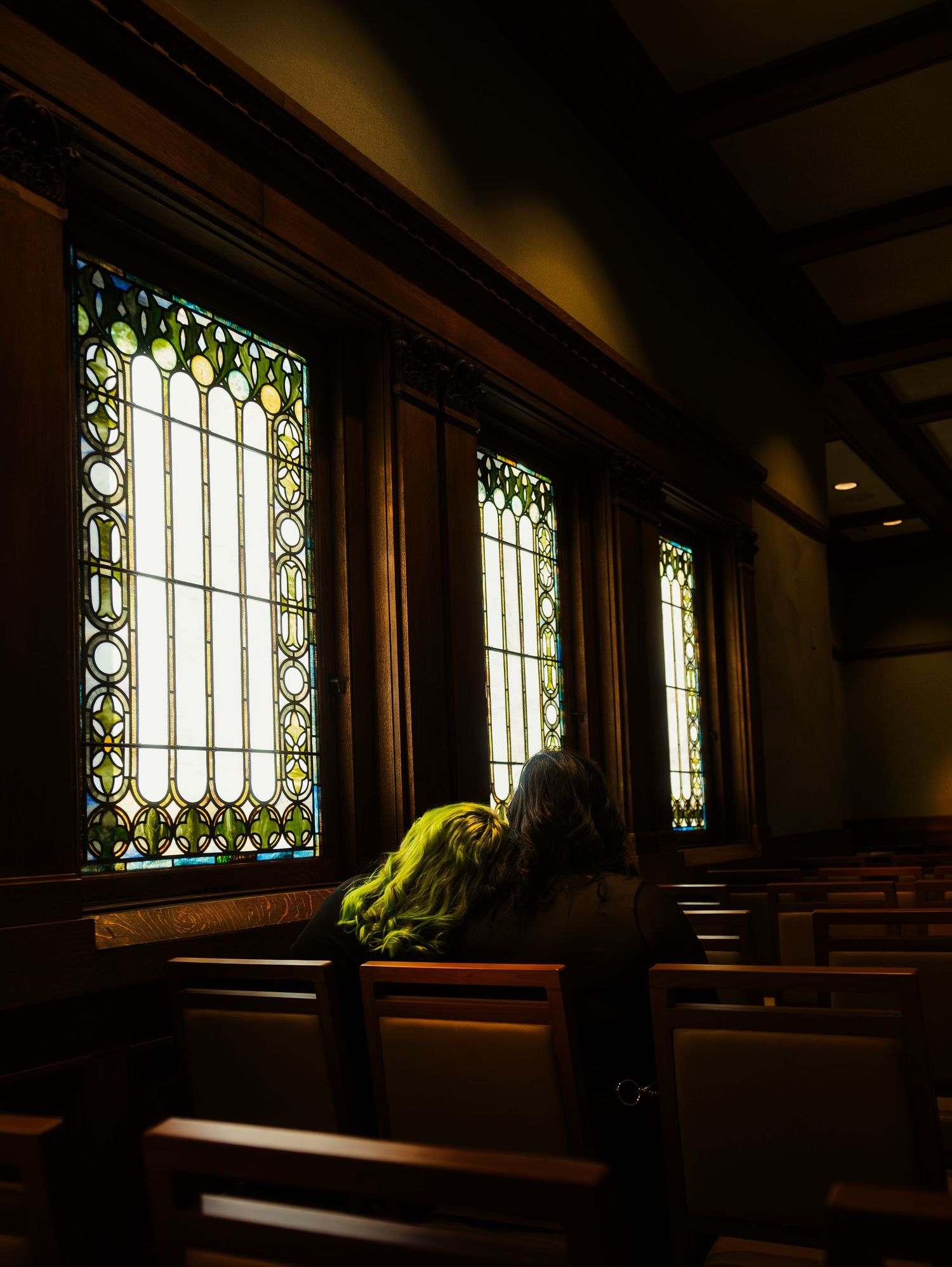 Sunlight streams through ornate stained glass windows in a dark church interior with wooden pews.