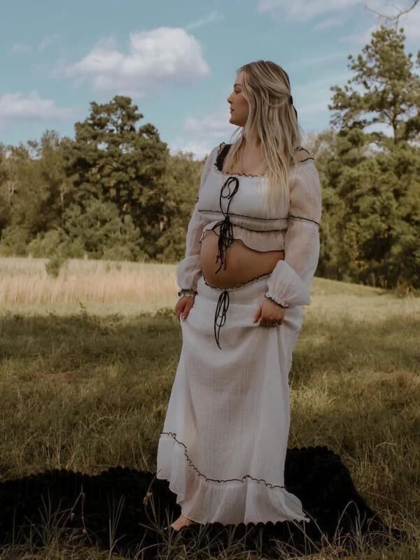 A person in a romantic cream-colored prairie dress with ruffles stands in a sunlit meadow surrounded by pine trees.