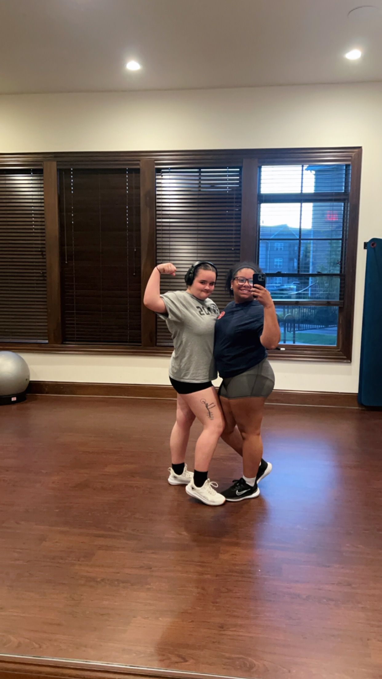 Two friends taking a mirror selfie while posing together in a workout room with wooden floors.