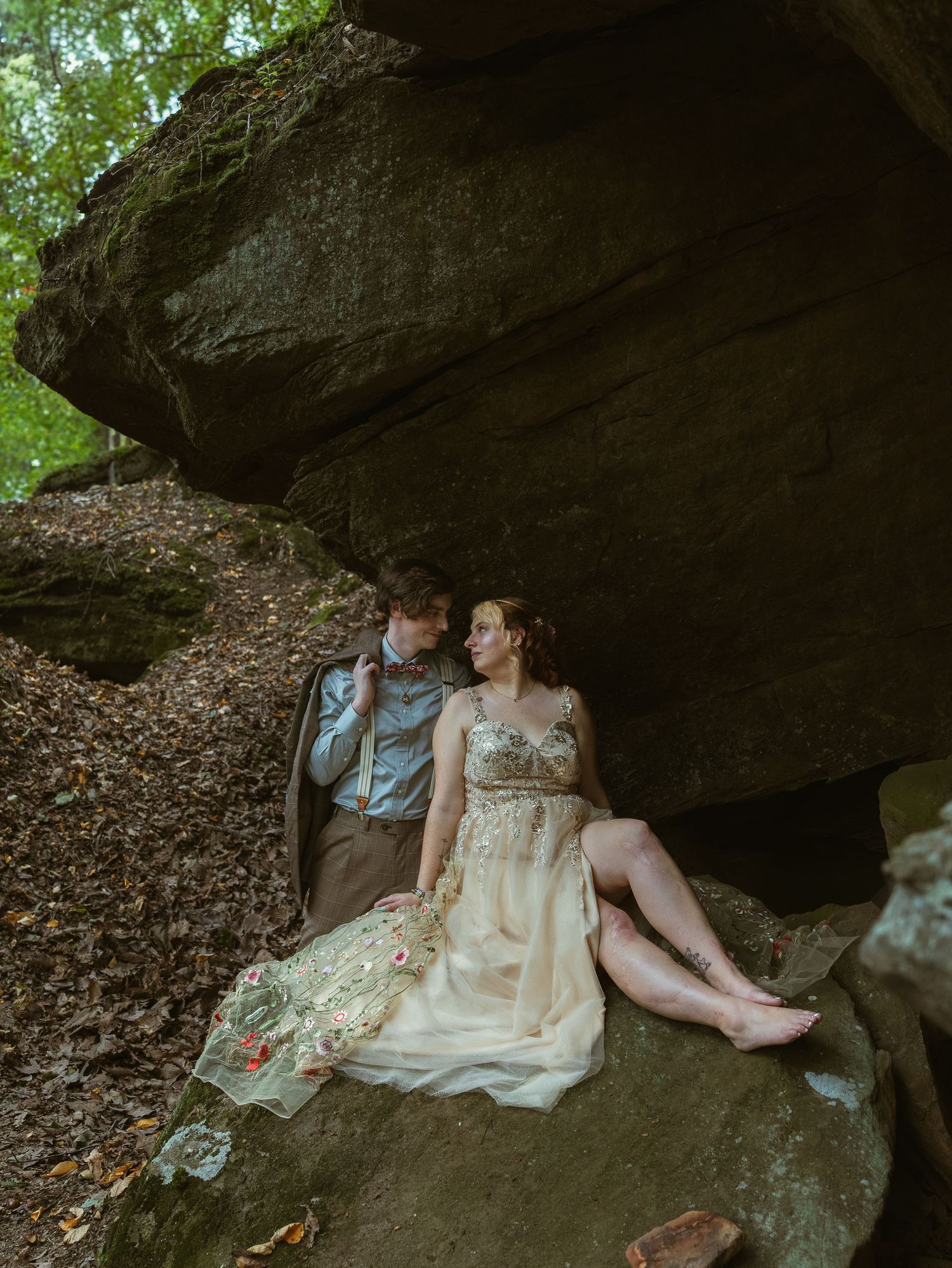 A couple in wedding attire rests intimately against a large rock formation in a forest setting.