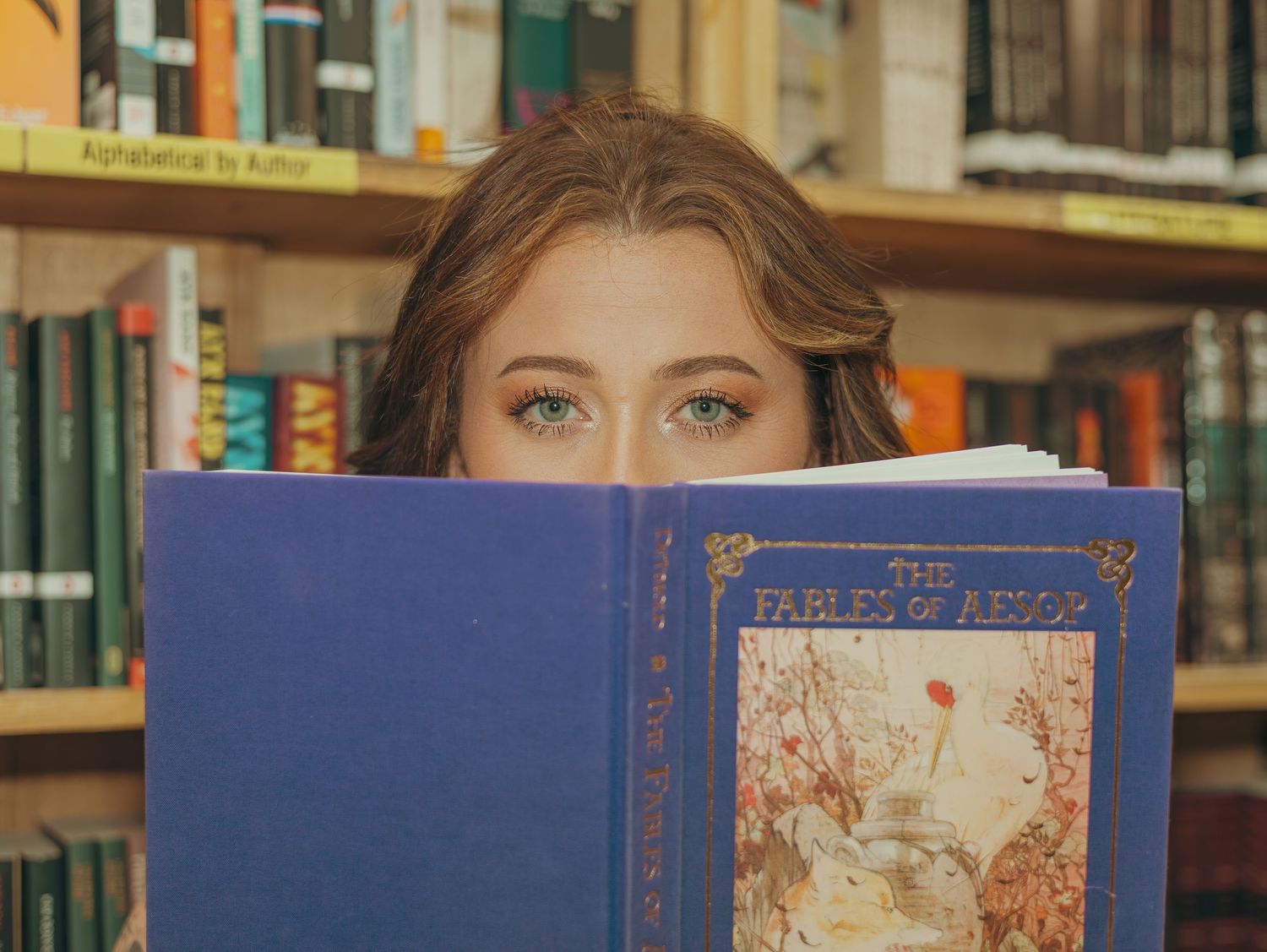 Someone holds up a blue vintage book in front of their face while standing in front of a bookshelf in a library or bookstore.