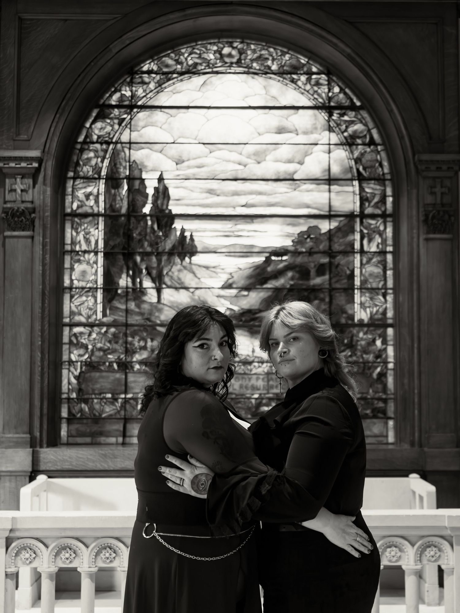Two women embrace before a stained glass window in a black and white portrait photo.