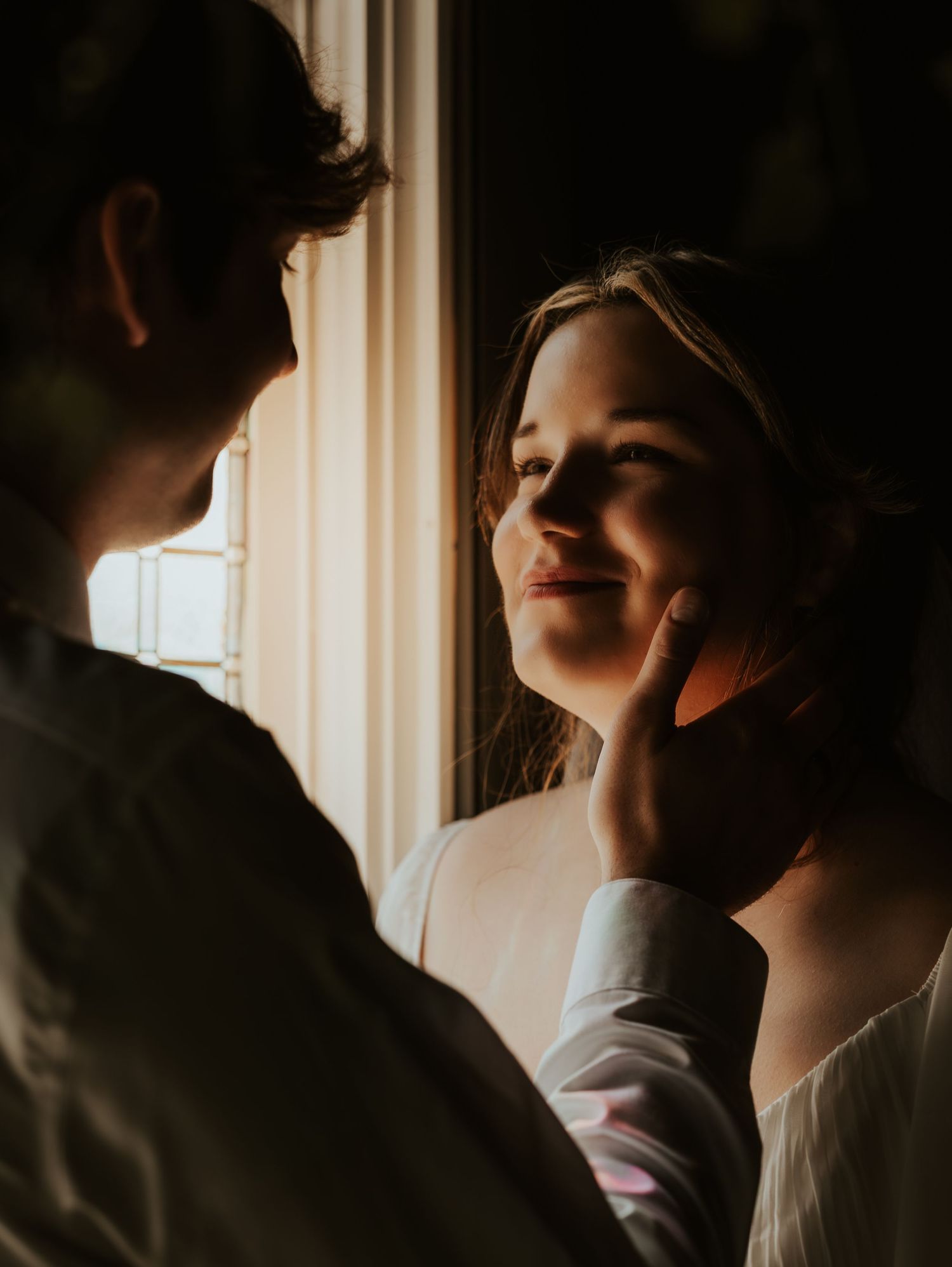 A couple share an intimate moment by a sunlit window creating a romantic silhouette in moody lighting.