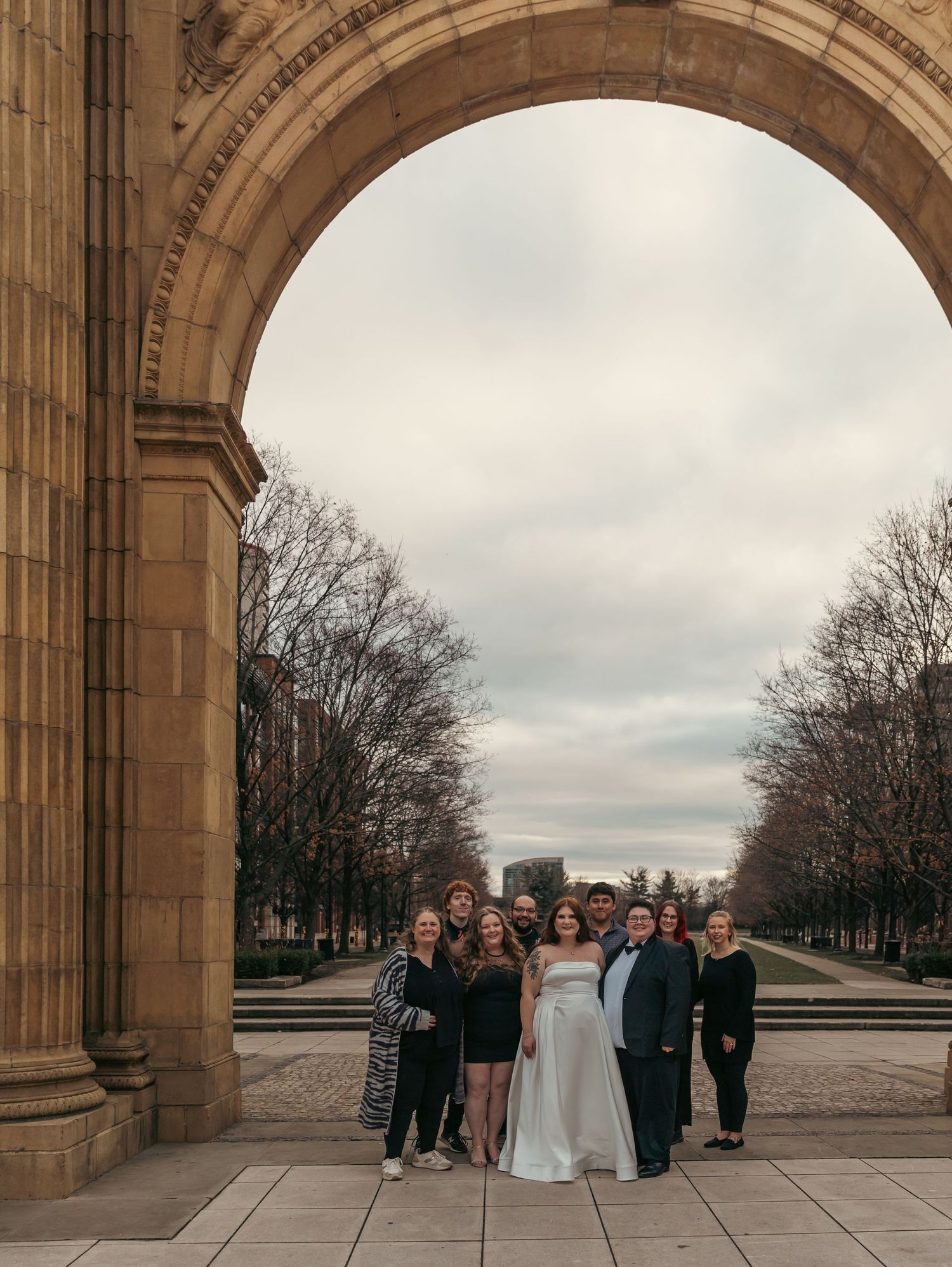 Wedding party gathered under large stone archway on overcast day.
