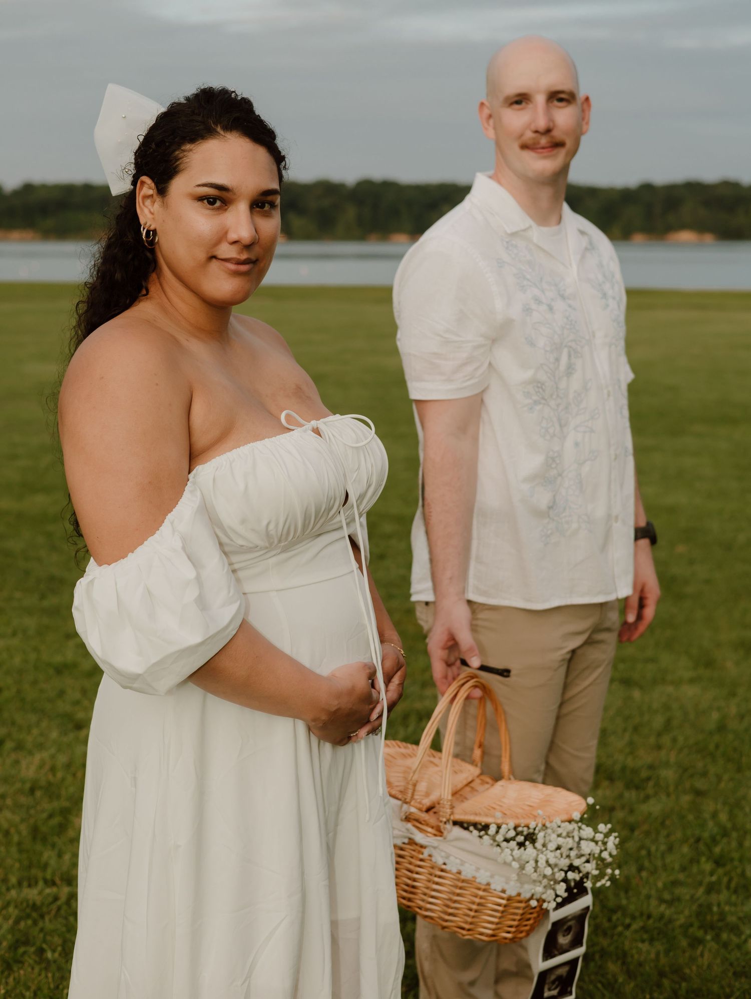 A couple in white and beige attire poses together in a grassy field with a woven basket of flowers.
