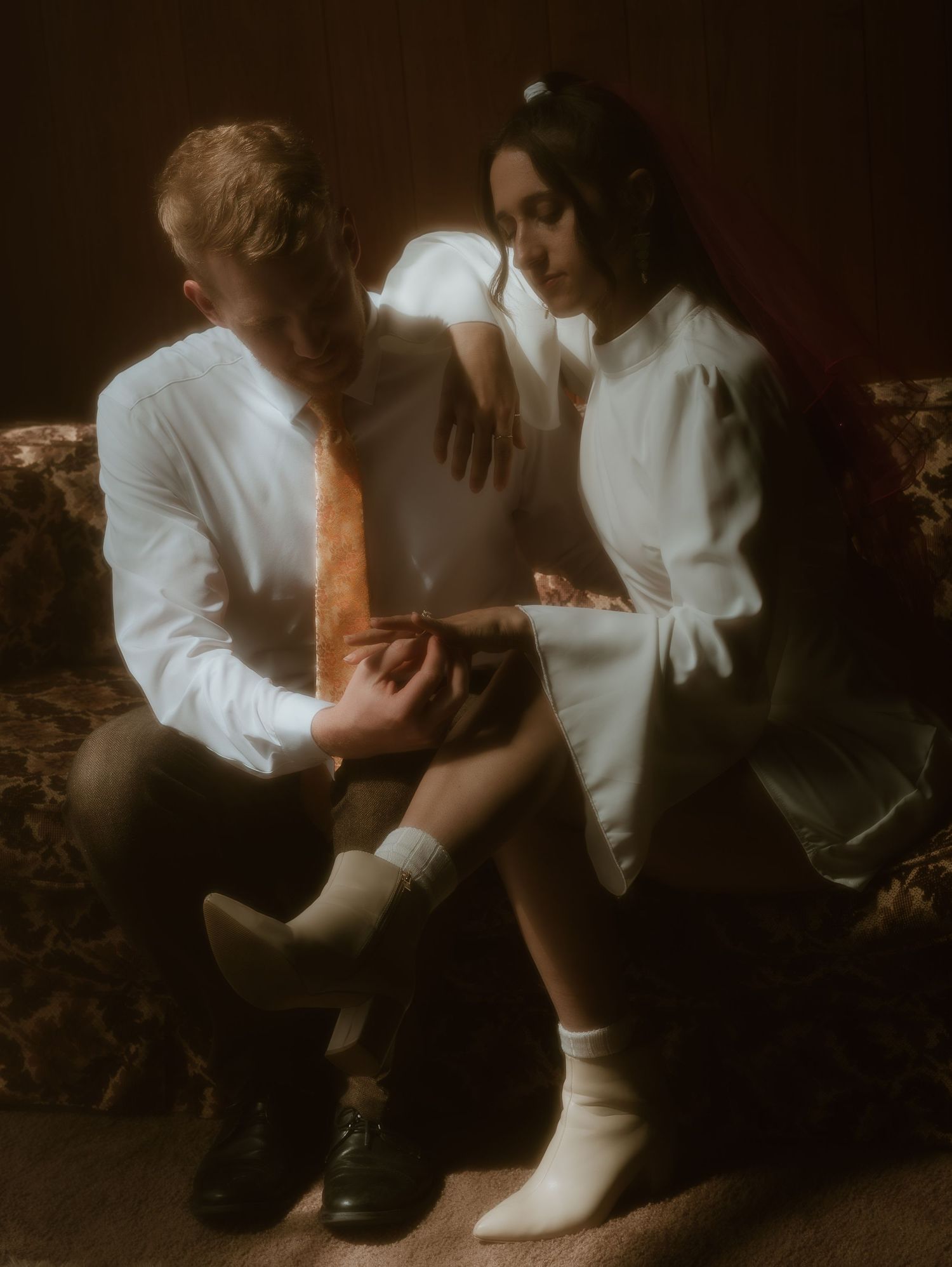 Two people in white clothing sit together on a dark wooden floor in dramatic moody lighting.