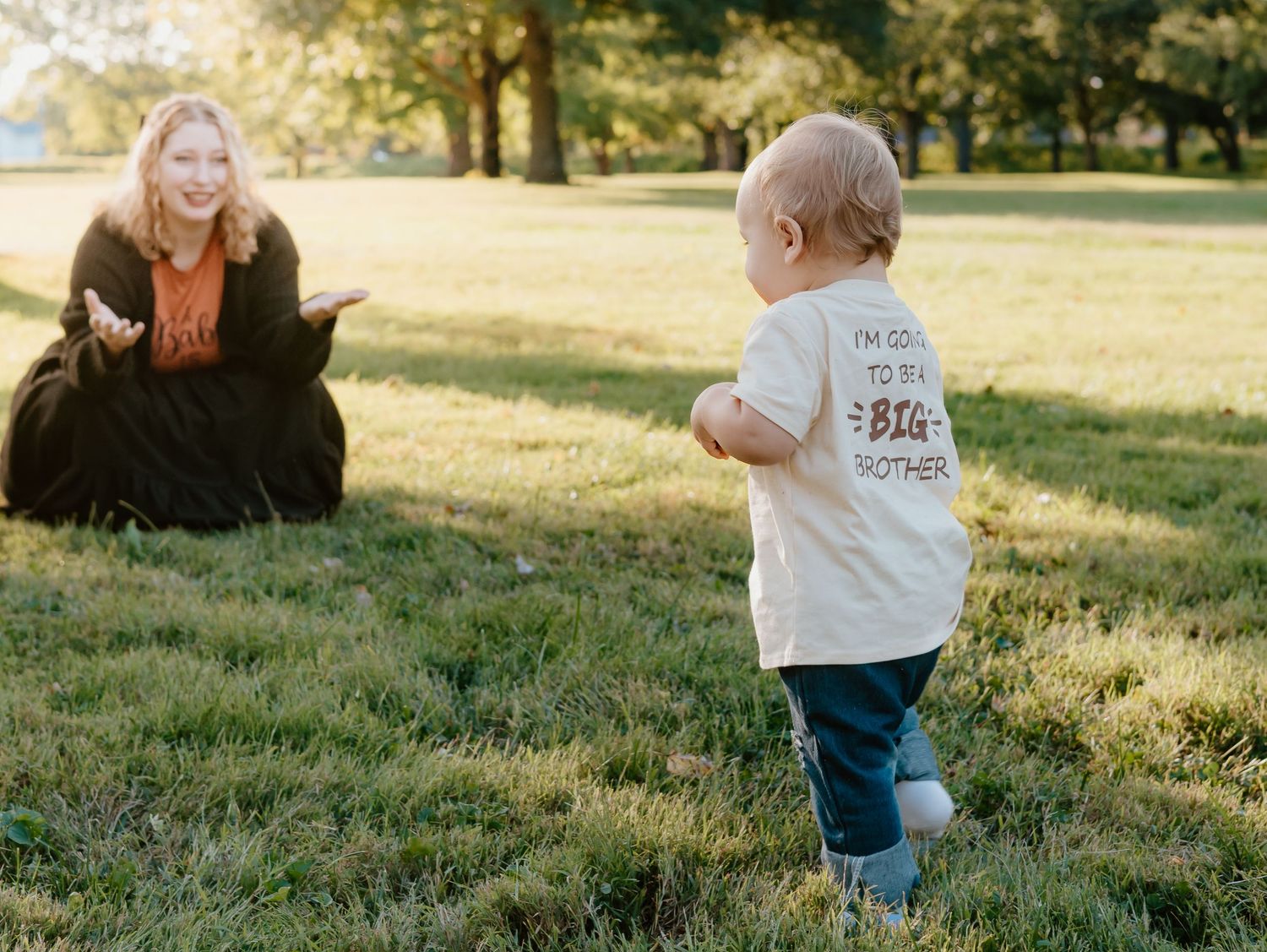 A toddler in jeans and white shirt runs through a sunlit grassy field during golden hour.