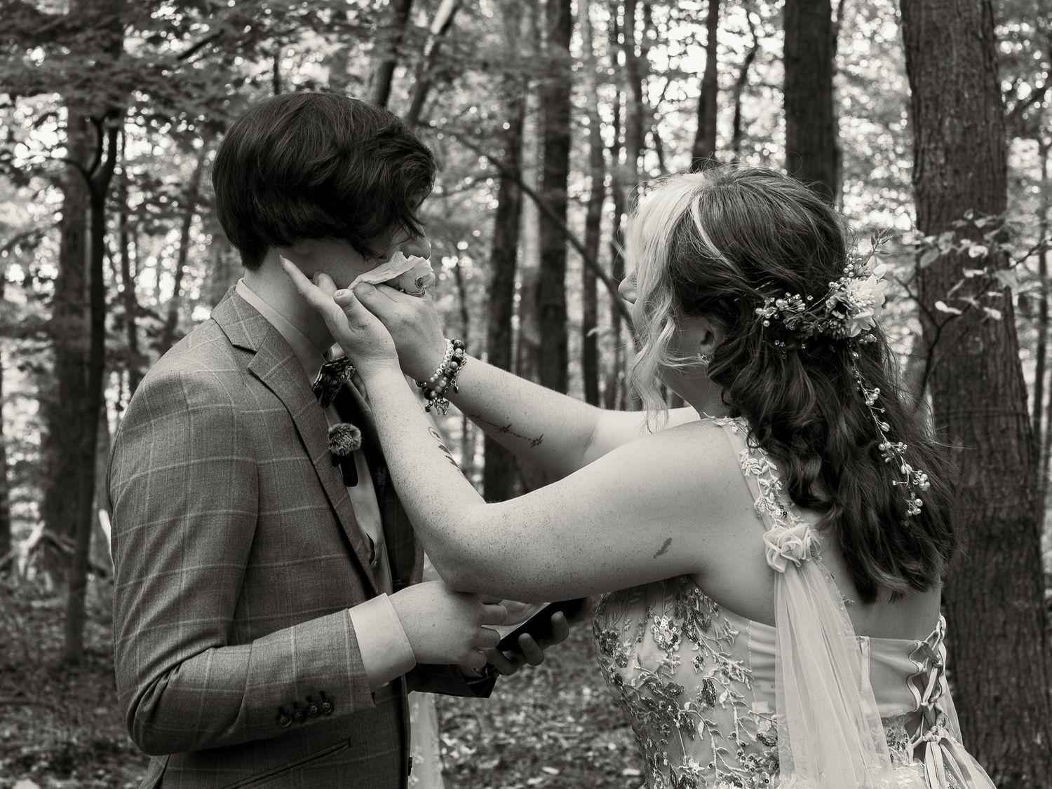 A bride and groom share an intimate moment in a wooded forest setting, captured in black and white photography.