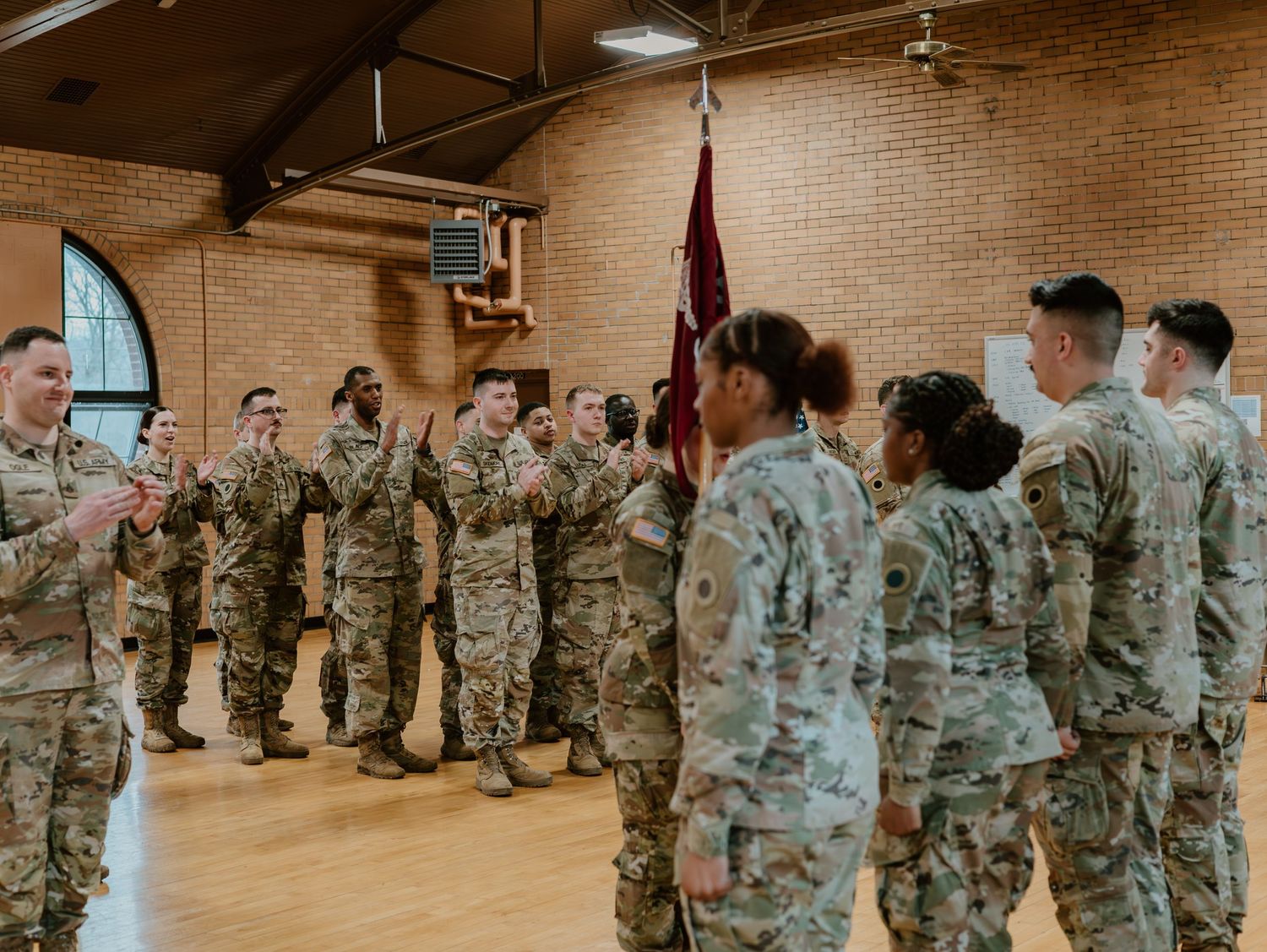 Soldiers in camouflage fatigues assemble in two rows inside a building with exposed brick walls and arched windows.