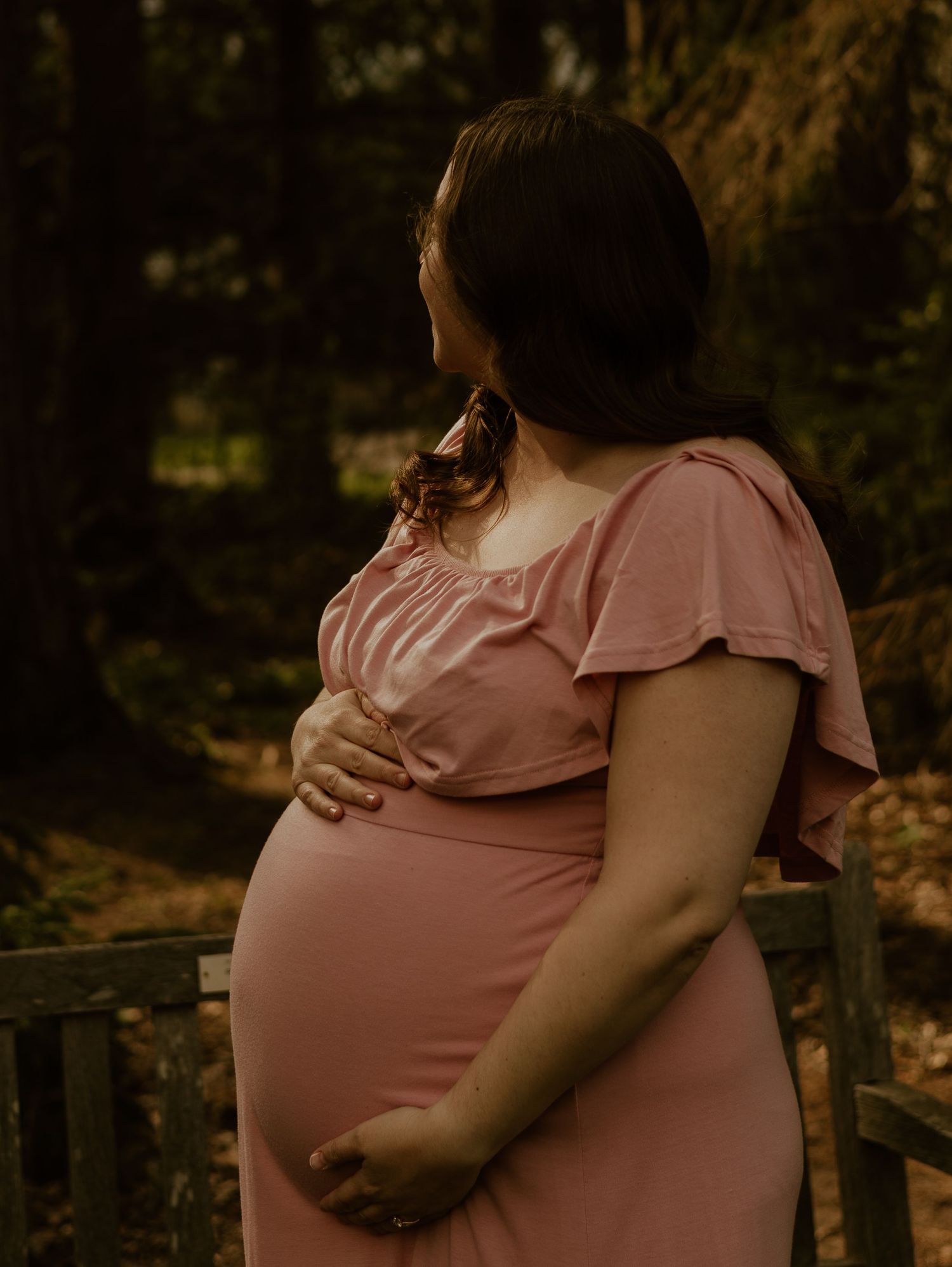 A pregnant woman in a pink dress stands in a wooded area during golden hour, cradling her baby bump.