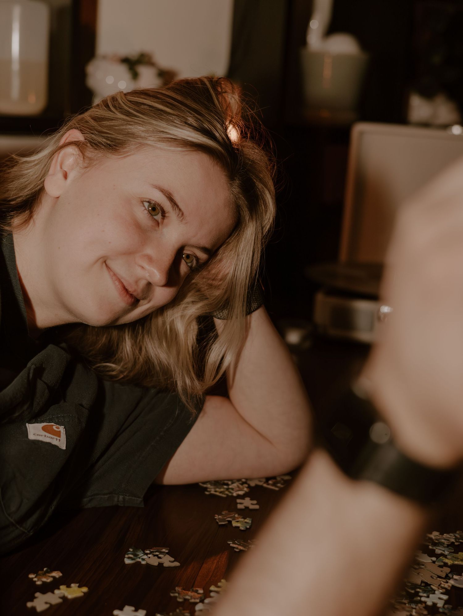 Someone lies on a table with scattered puzzle pieces in a moody, dimly lit setting with warm tones.