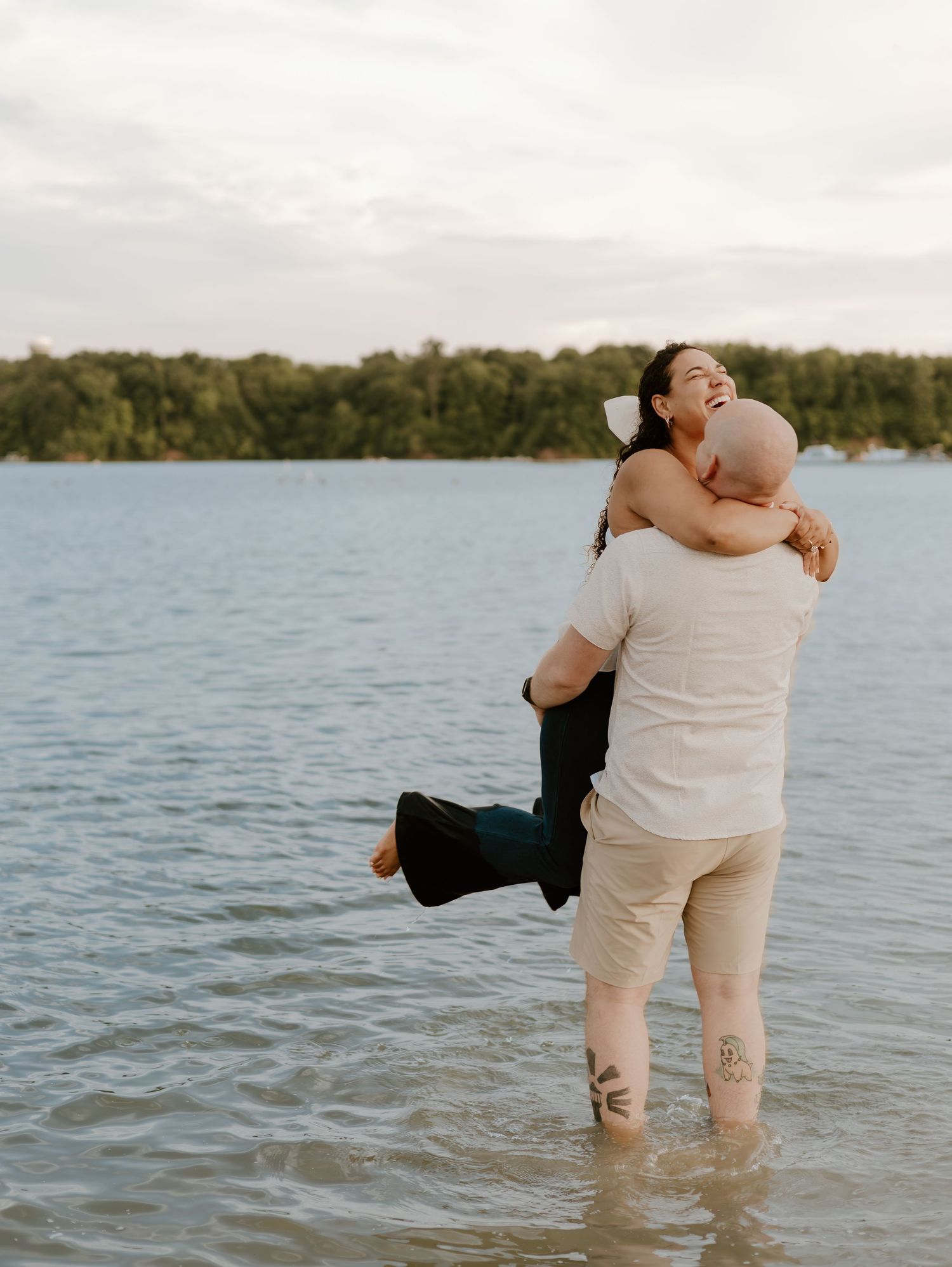 A couple embraces while standing in shallow lake water at sunset with trees visible along the shoreline.