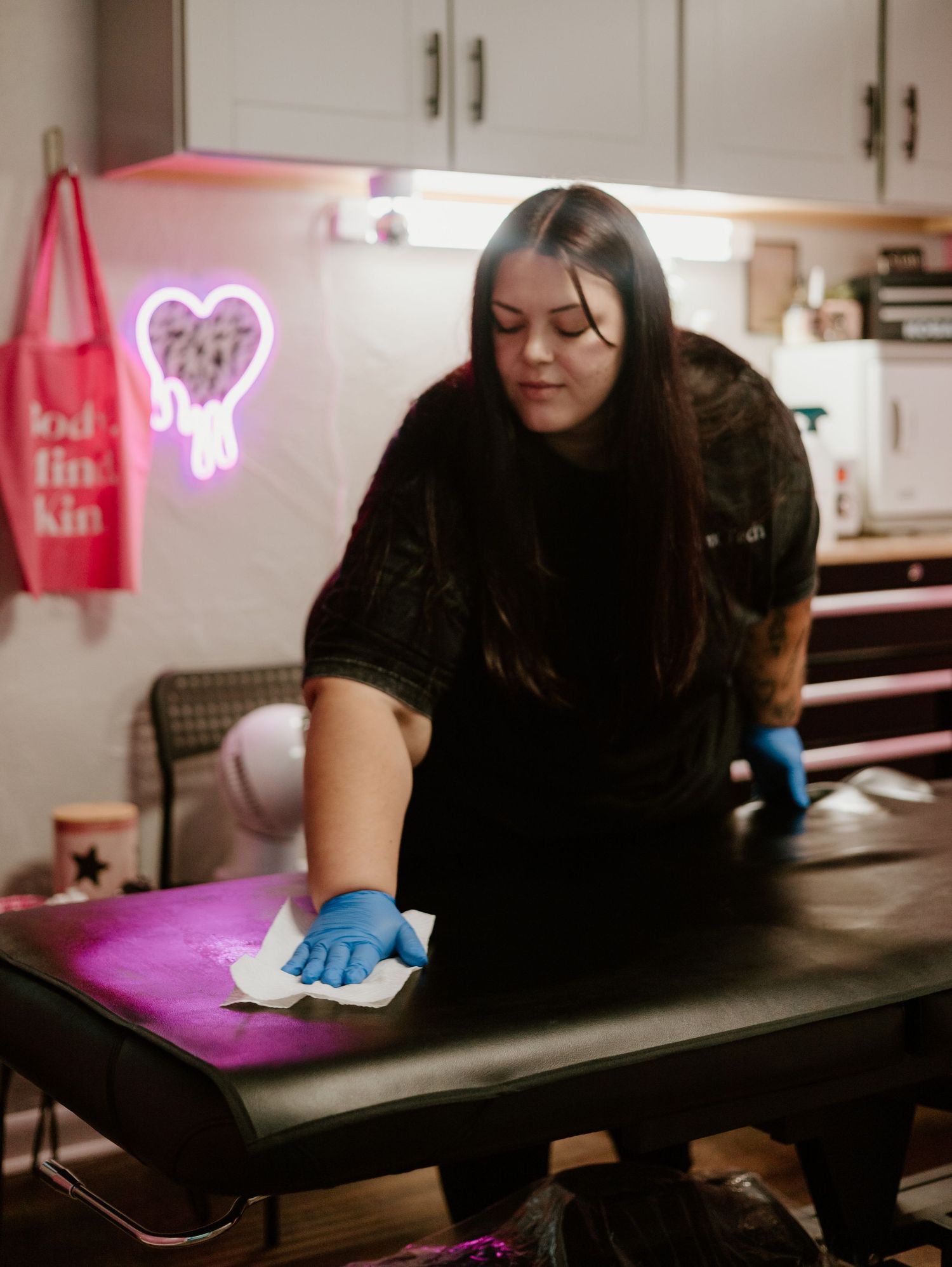 Someone wearing black clothing and blue gloves wipes down a table surface with a neon heart sign glowing in the background.