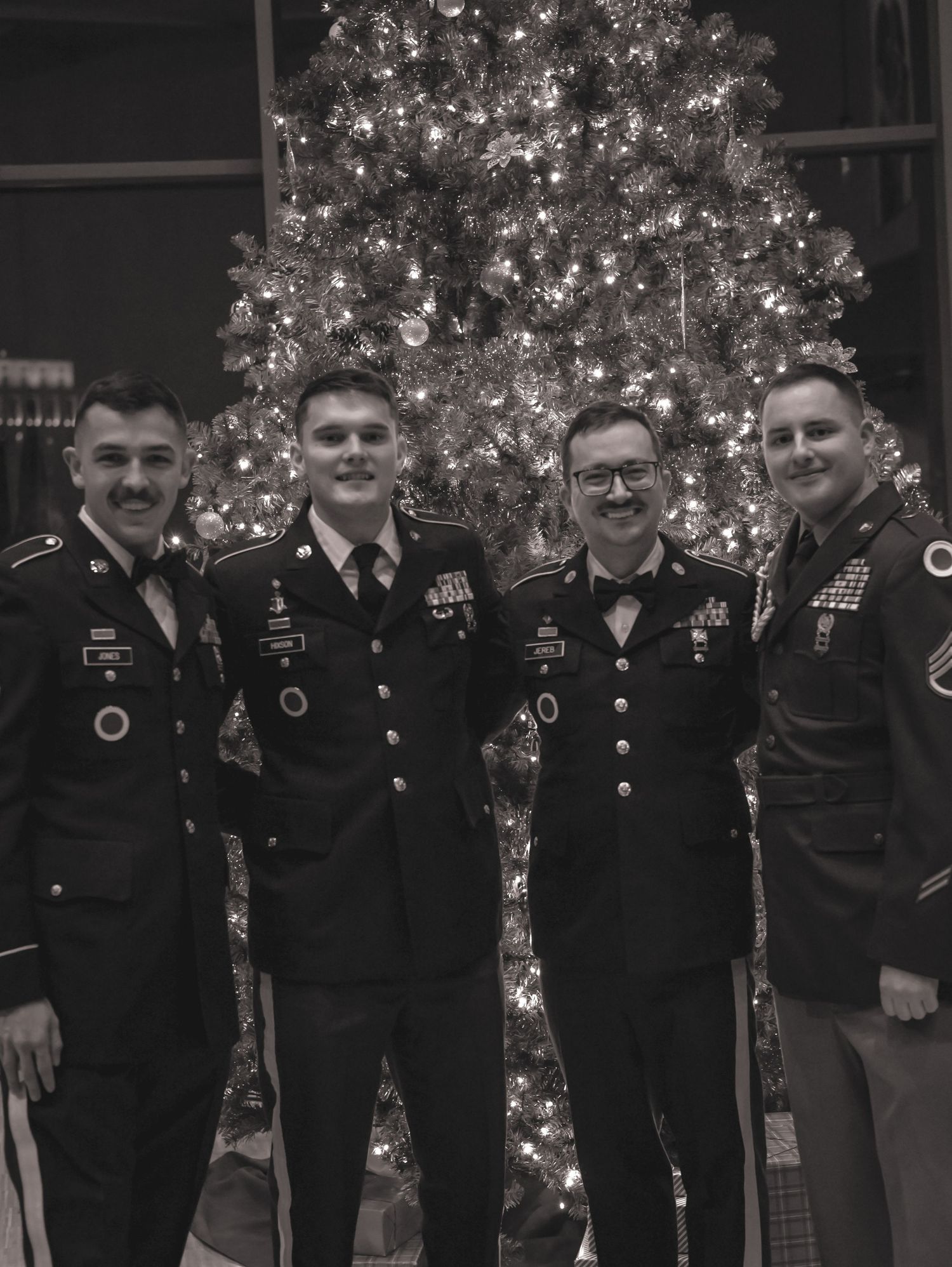 Four service members in dress uniforms pose in front of a glowing Christmas tree.