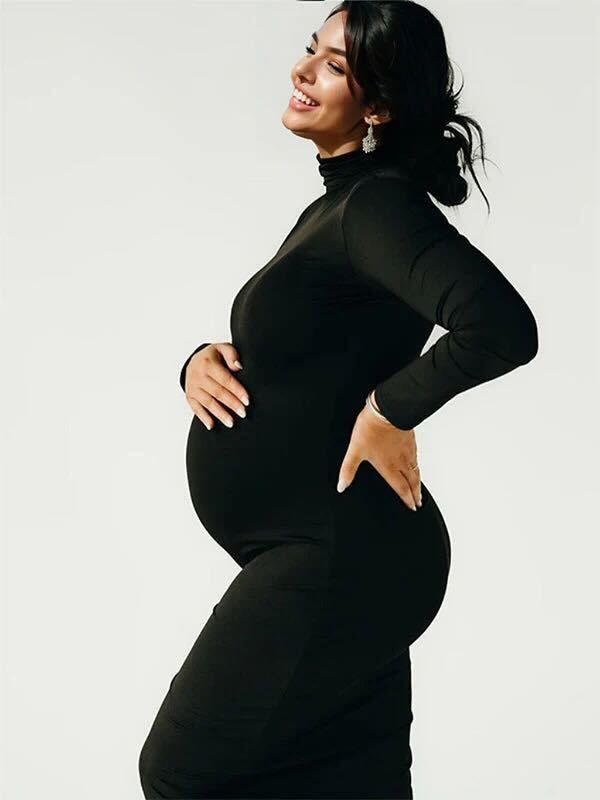 Side view of a pregnant woman wearing a black turtleneck dress posing gracefully against a white background.