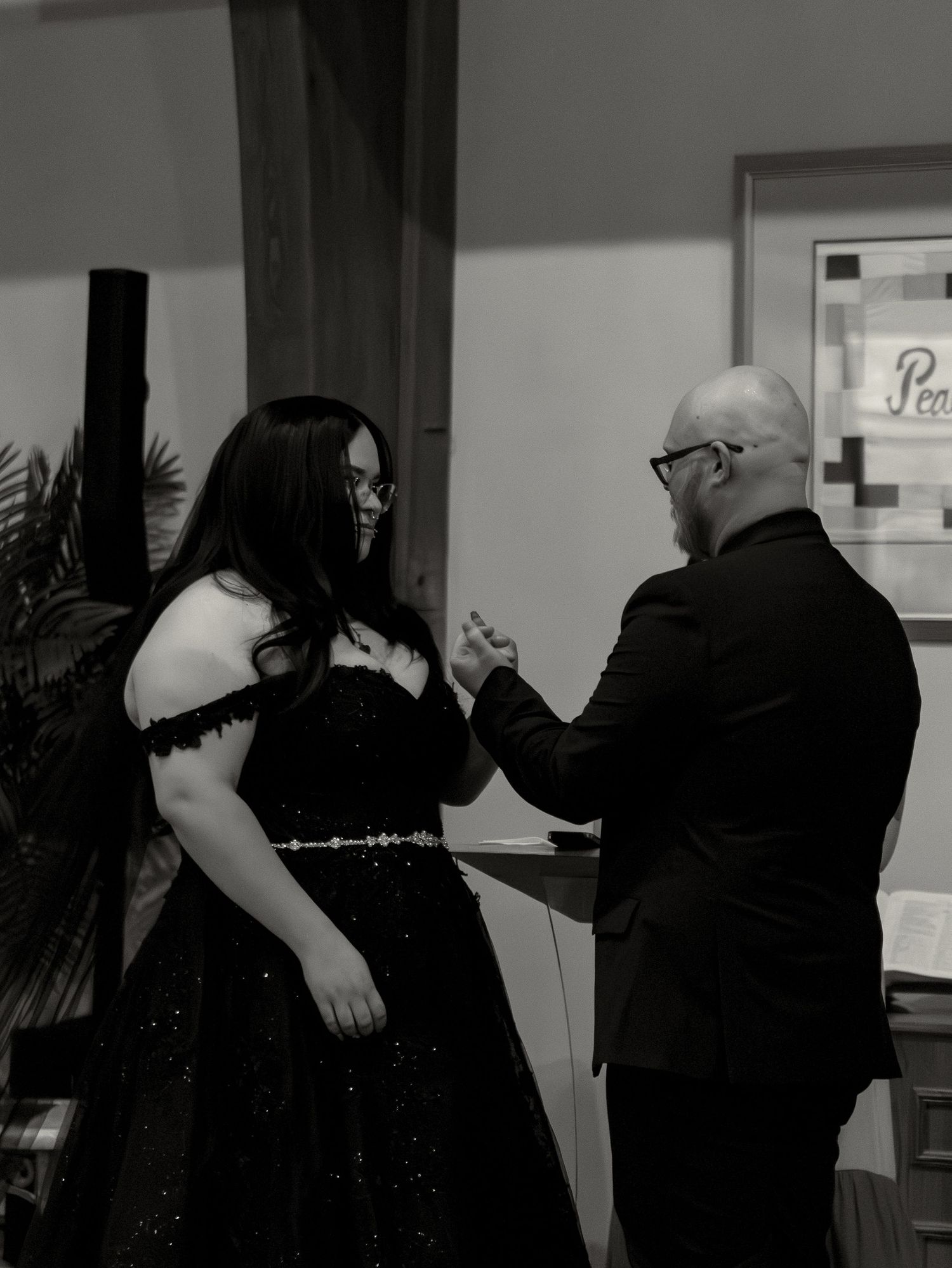 Black and white photo of a couple in formal black attire holding hands at an elegant indoor event.
