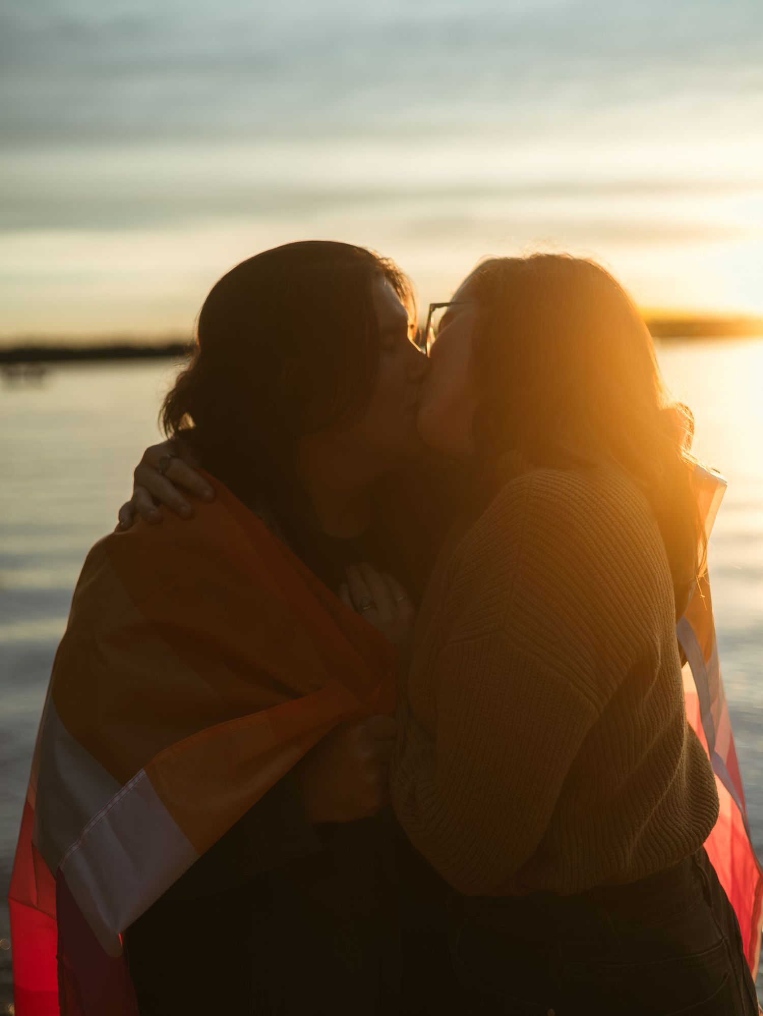 Two women sharing a kiss wrapped in an LGBT pride flag at sunset by the water.