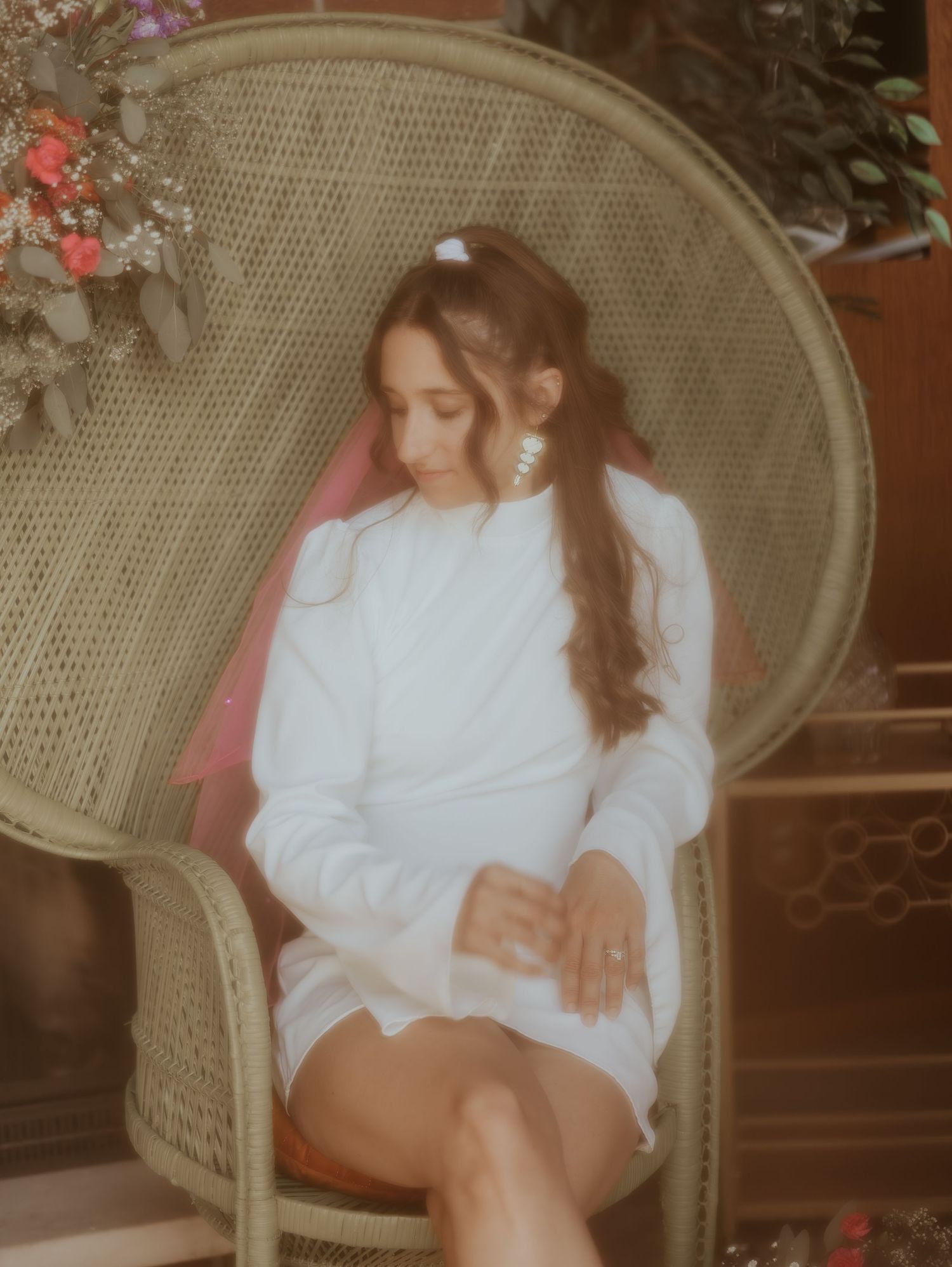 A bride in a white sweater poses elegantly on a vintage wicker chair with floral decor.