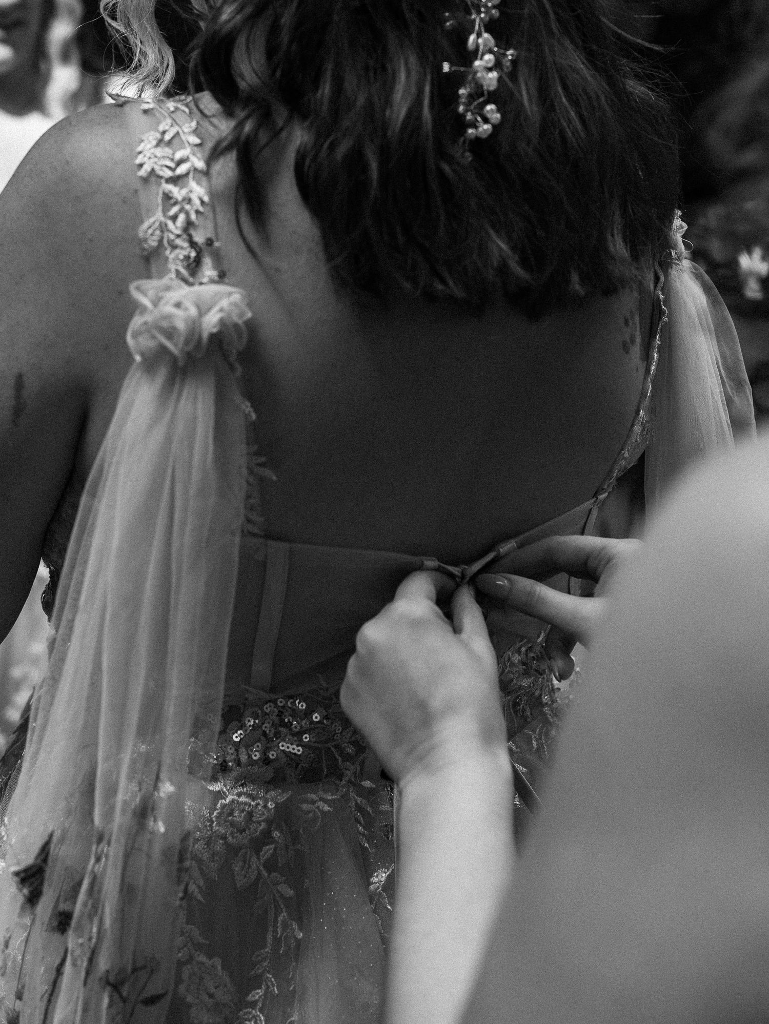 Black and white close-up of hands lacing up an ornate wedding dress with delicate floral details.