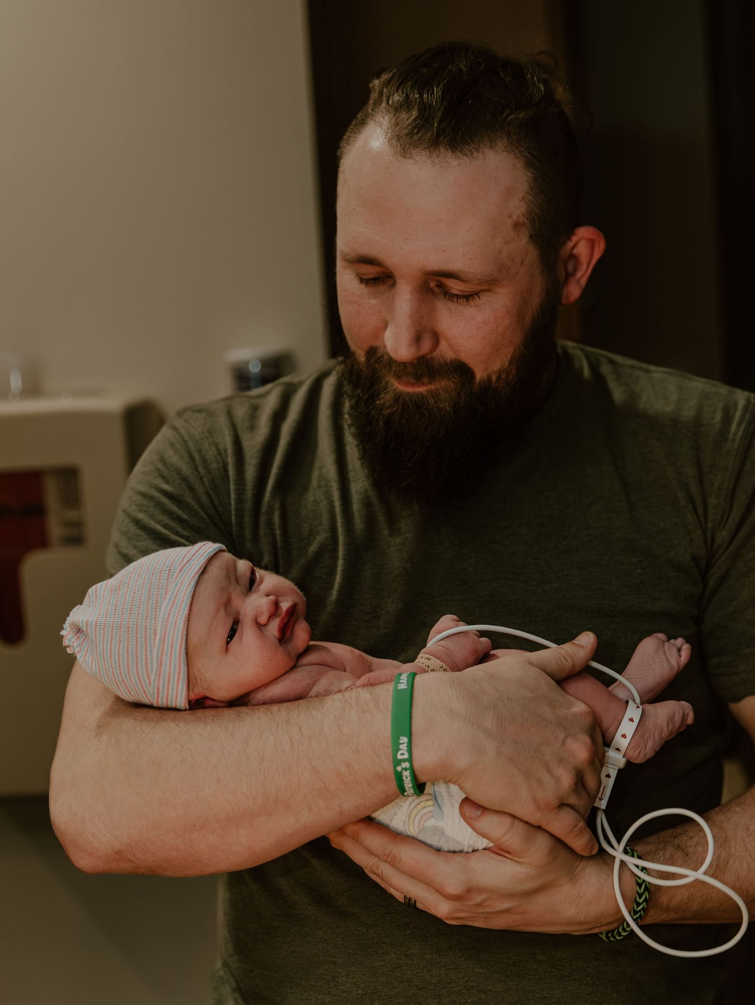 A bearded man in a green shirt tenderly holds a newborn baby wearing a hospital cap in what appears to be a medical setting.