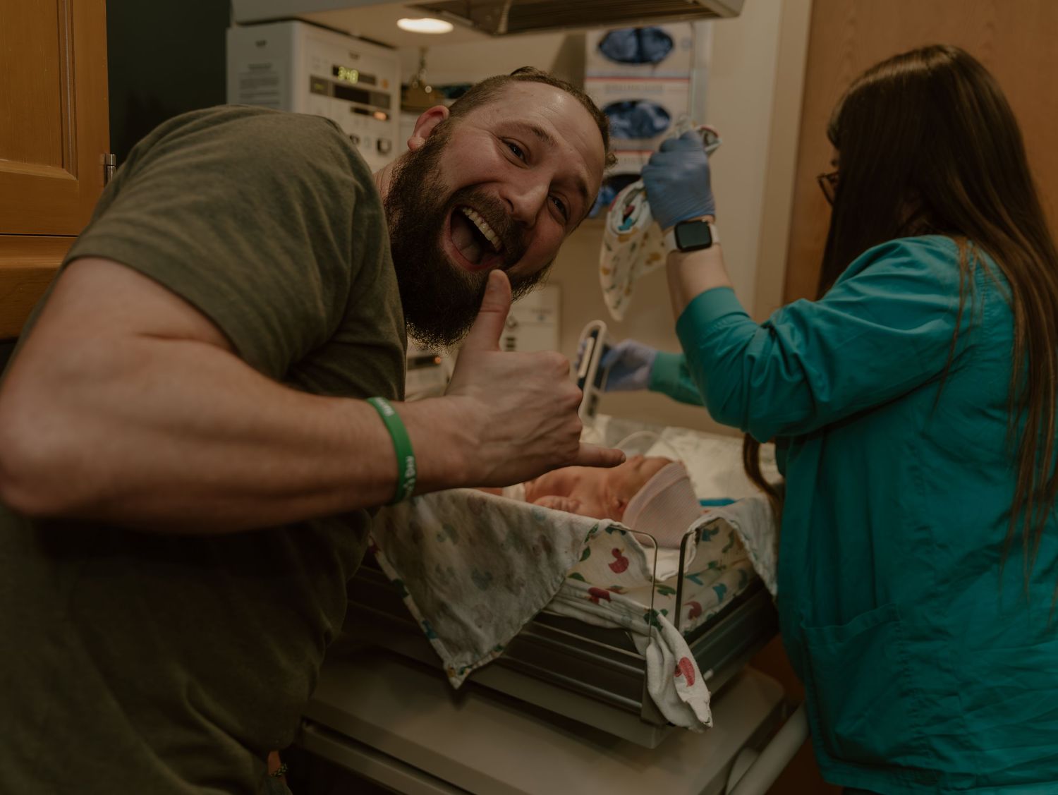 Excited man smiling broadly next to a hospital newborn bassinet while a nurse in teal scrubs works nearby.