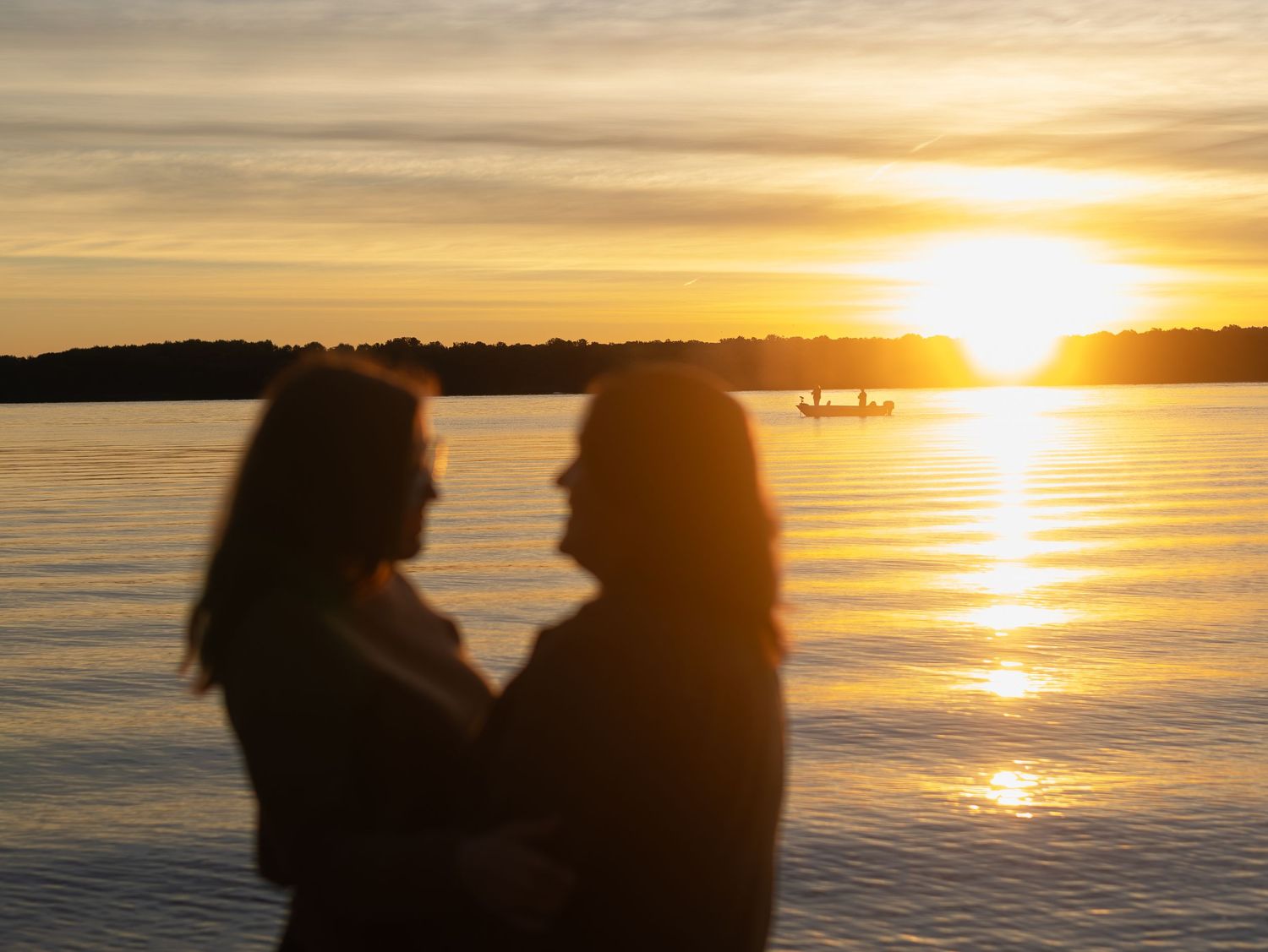Two silhouetted figures face each other romantically at sunset over a calm lake with golden reflections.
