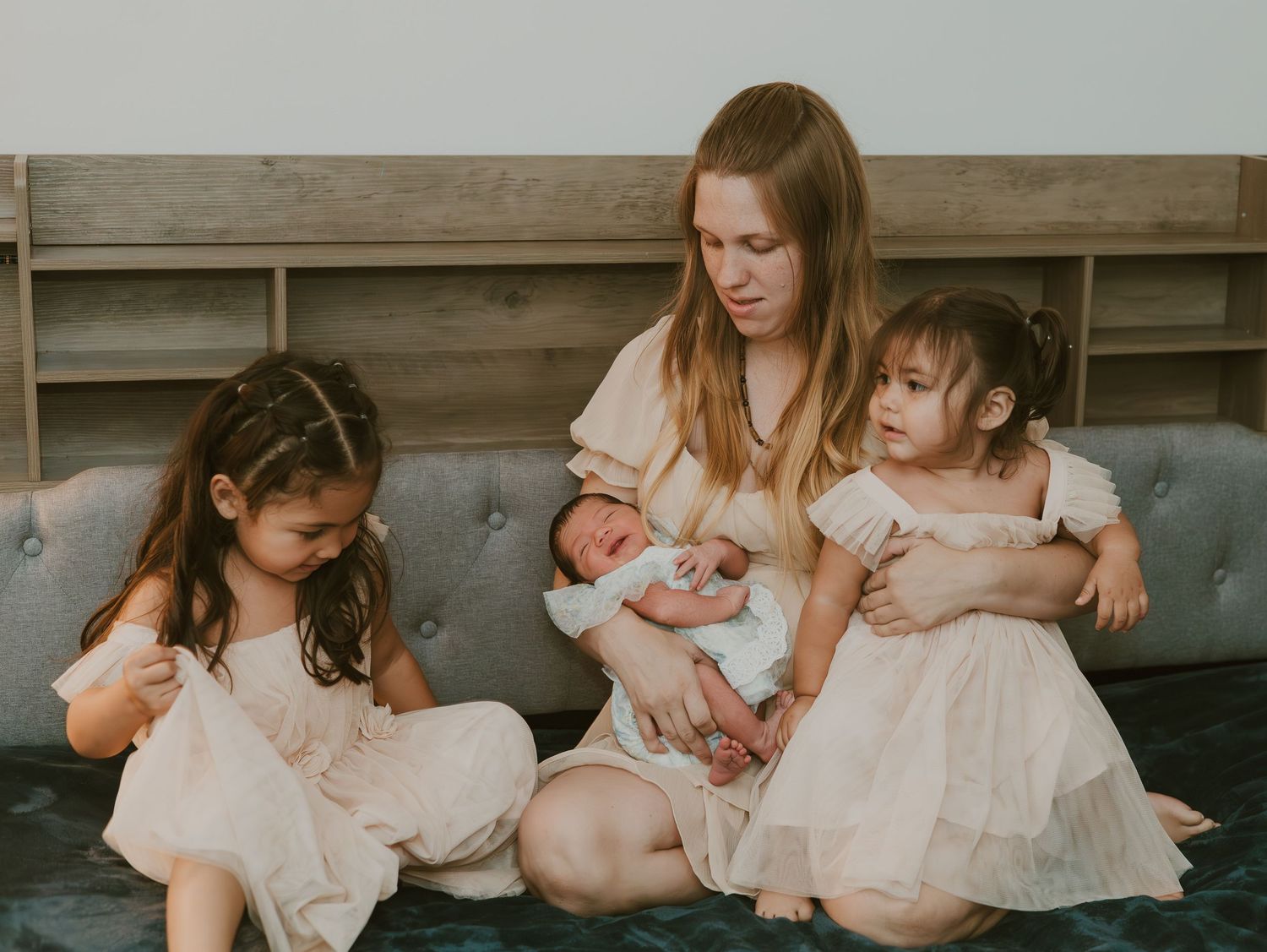 Family wearing soft pink and cream colored outfits sits together on wooden floor during indoor photography session.