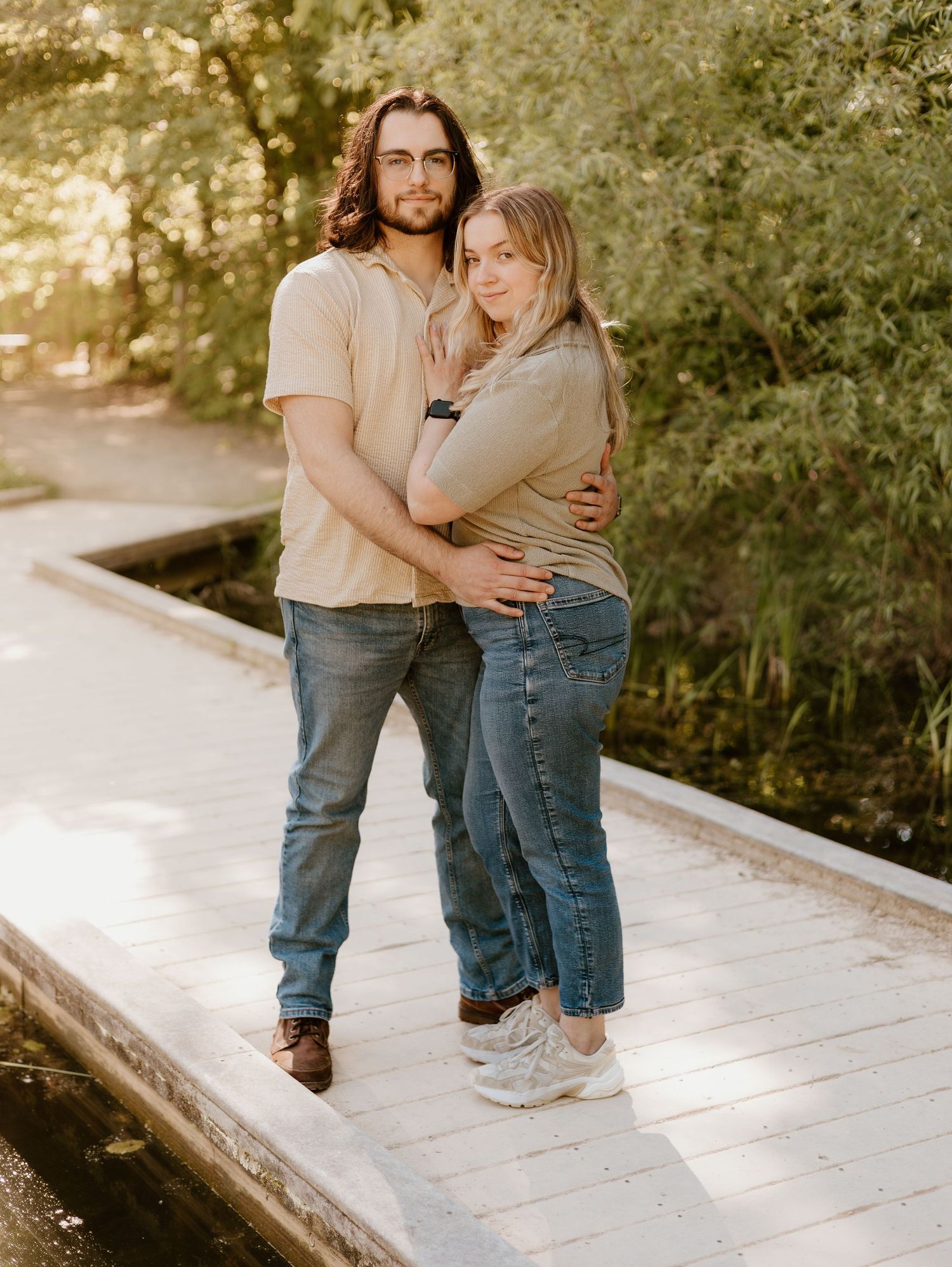 A couple stands close together on concrete steps surrounded by lush greenery in warm sunlight.