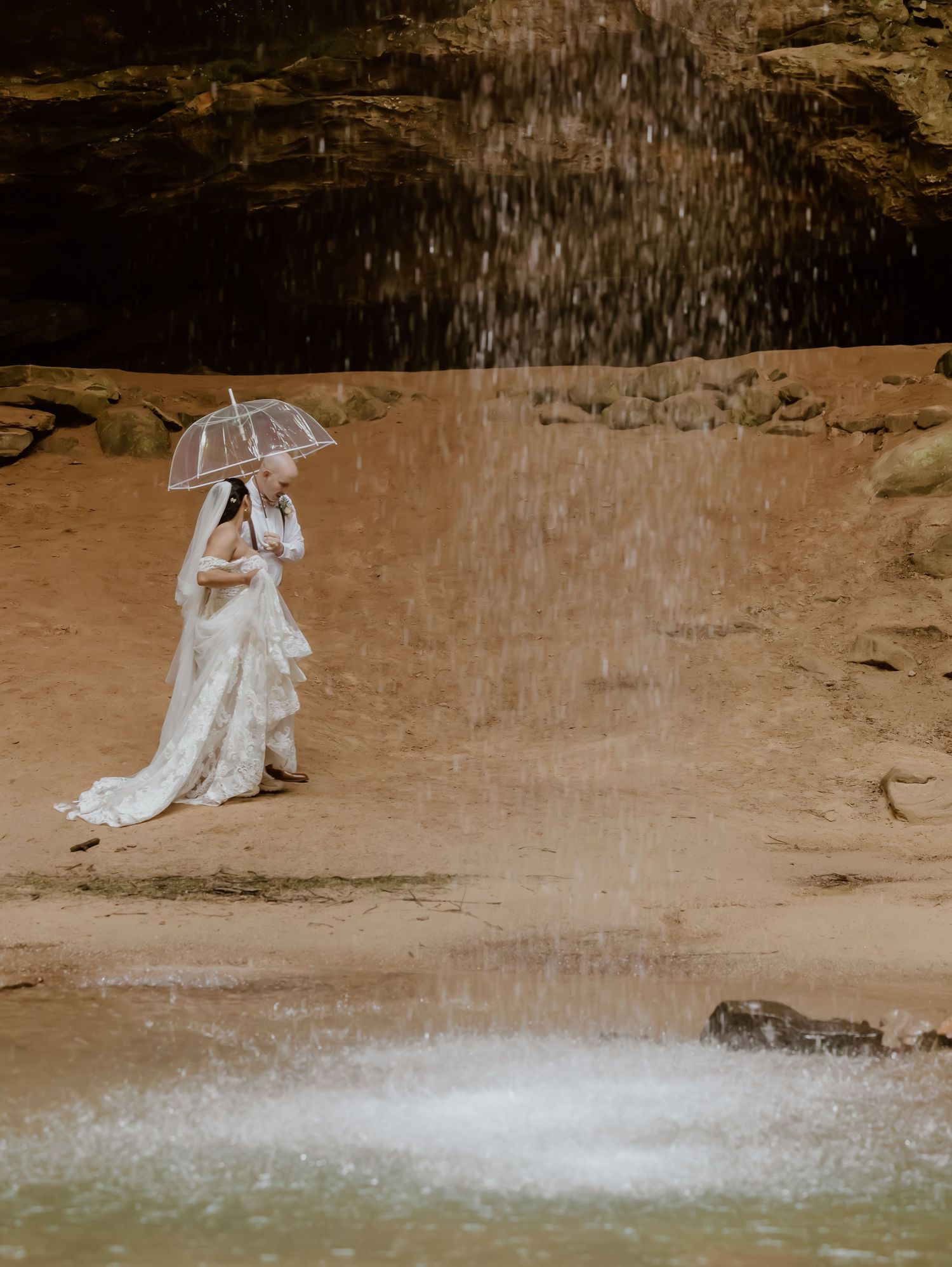 Wedding couple posing with clear umbrellas under a waterfall spray.