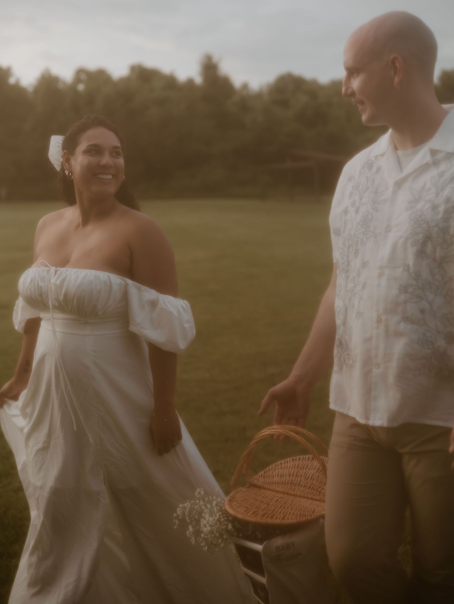 A couple in white outfits walking through a grassy field at sunset with a picnic basket.
