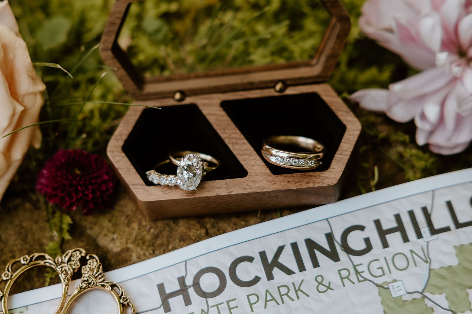 Wedding rings displayed in a wooden hexagonal box next to a Hockinghill sign surrounded by flowers and moss.