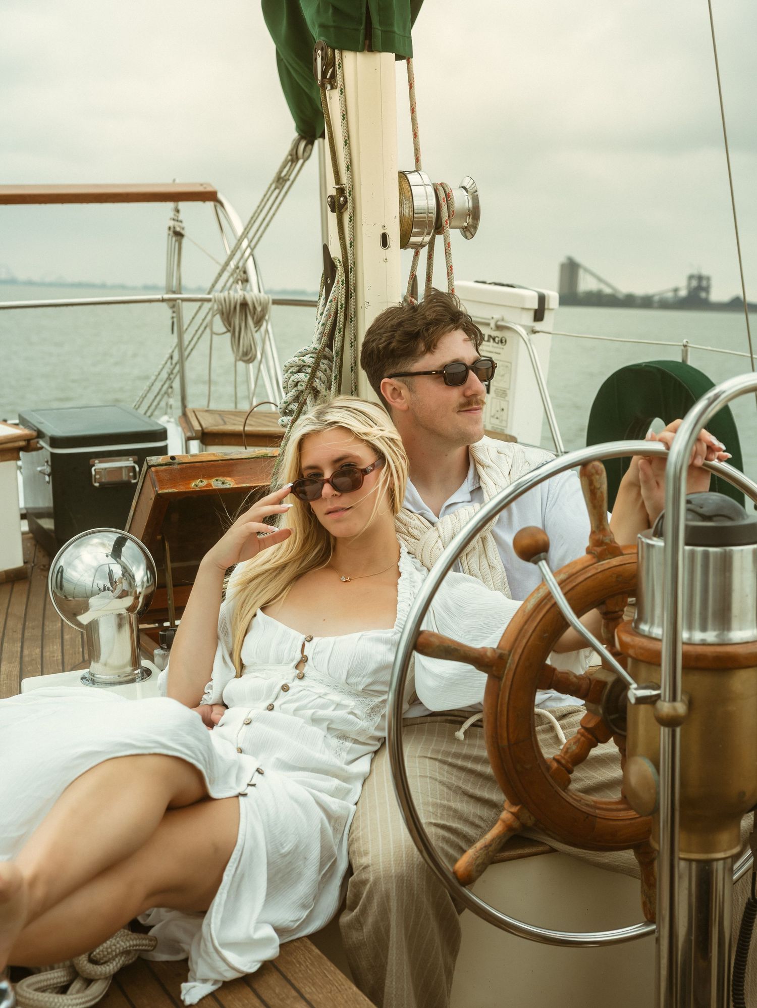 A couple in white outfits relaxes on a classic sailboat deck, with a bridge visible in the misty background.