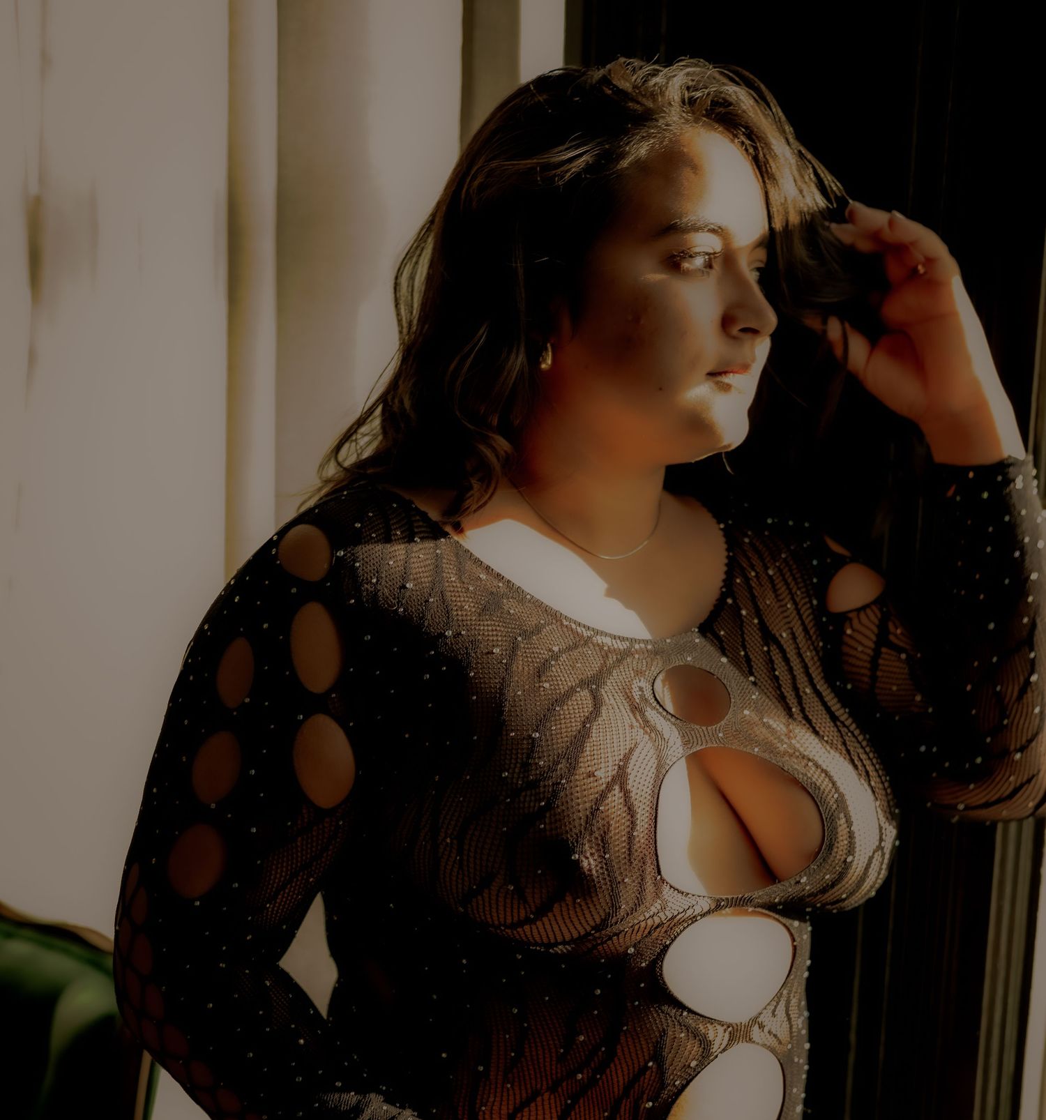 Person posing in black mesh bodysuit near window with dramatic lighting.